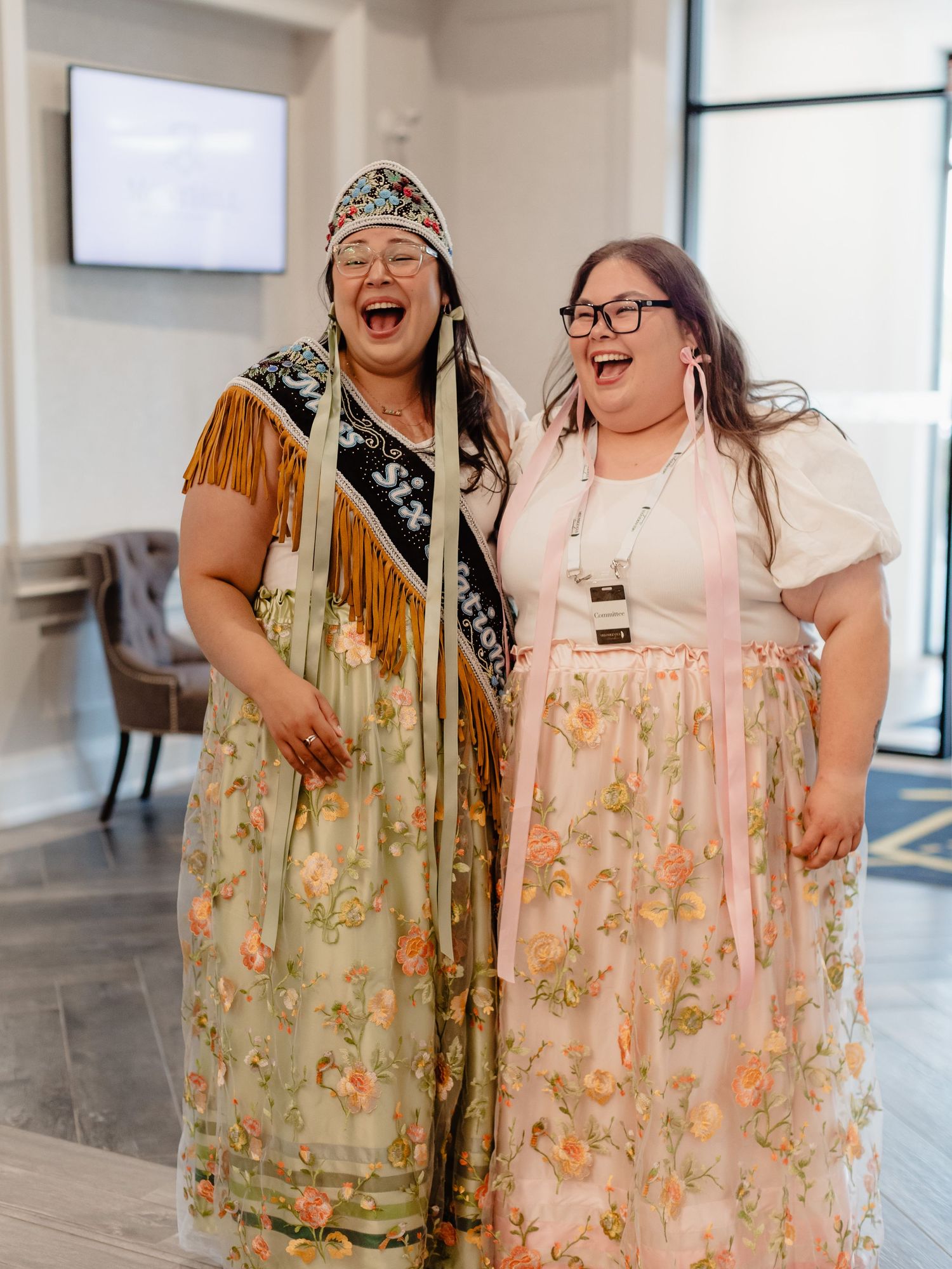 Two women in traditional Indigenous floral regalia laugh joyfully together indoors.