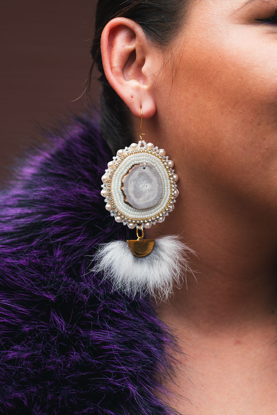 Woman wearing elegant oval geode earring with pearl border and white feather tassel, paired with purple fur coat.