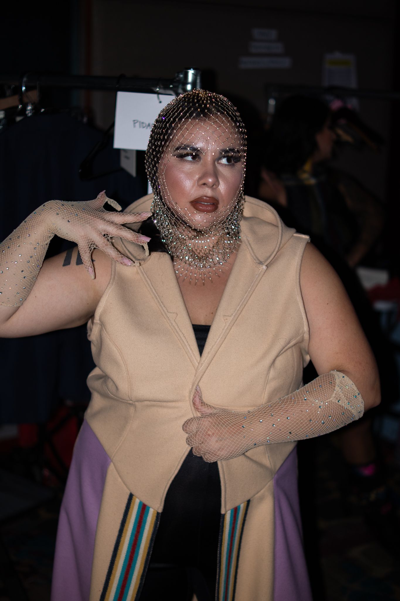 Model poses in beige vest with rhinestone mesh headpiece and gloves at a fashion show backstage.