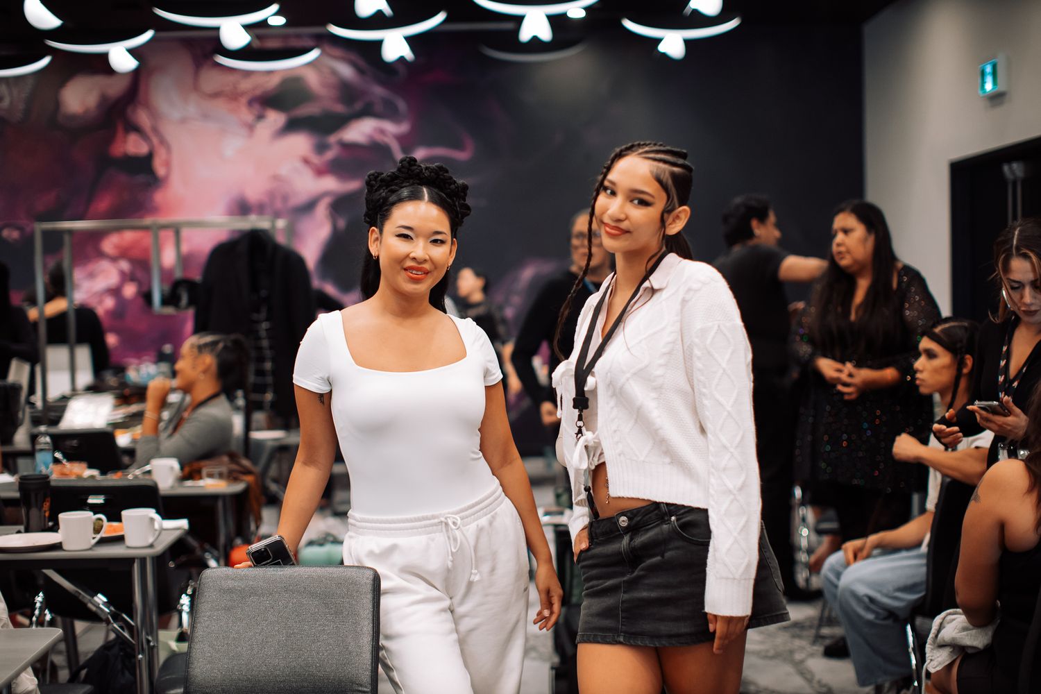 Two women pose together in a modern salon studio, one in white outfit and one in white shirt with black skirt.