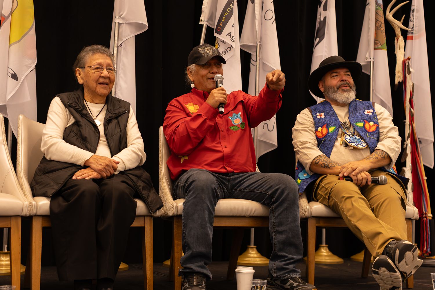 Three Native American elders sit on stage at a conference, one speaking into a microphone with tribal flags displayed behind them.