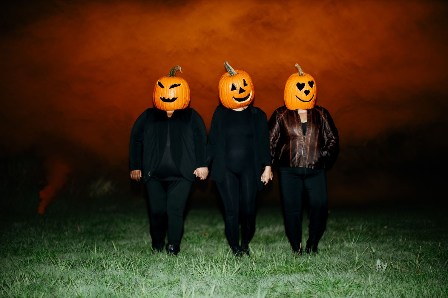 Group wearing pumpkin heads standing in a field at dusk with orange sky.