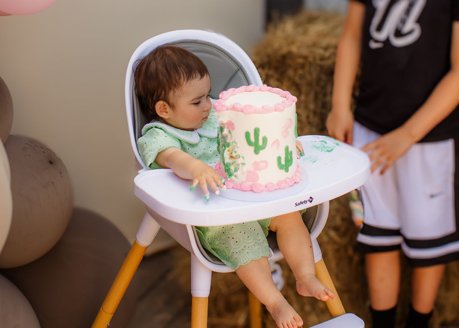 A baby in a high chair reaches for a pink cactus smash cake at a first birthday party.