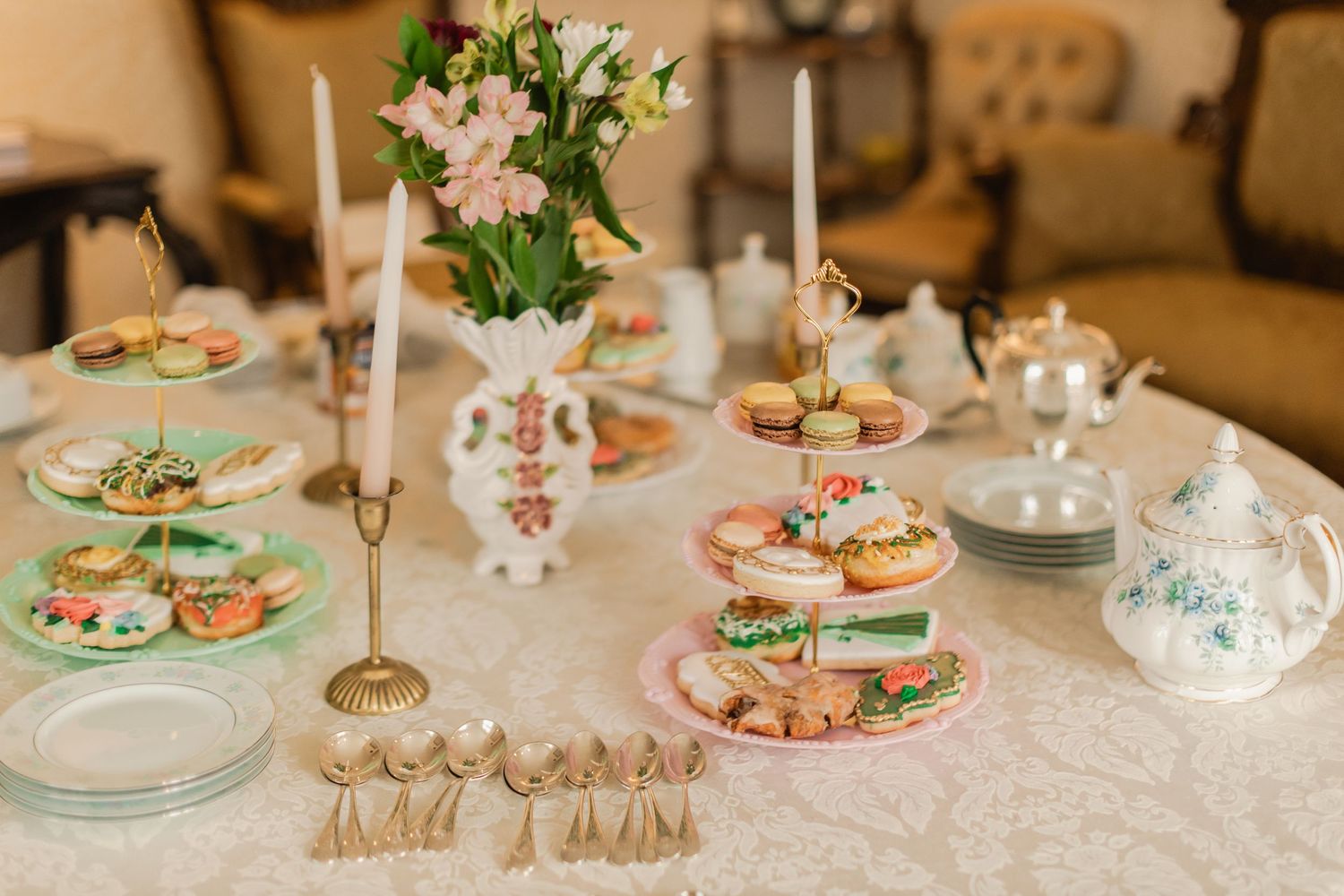 Elegant high tea table spread with tiered pastry stand, macarons, candles, fresh flowers, and fine china on a lace tablecloth.