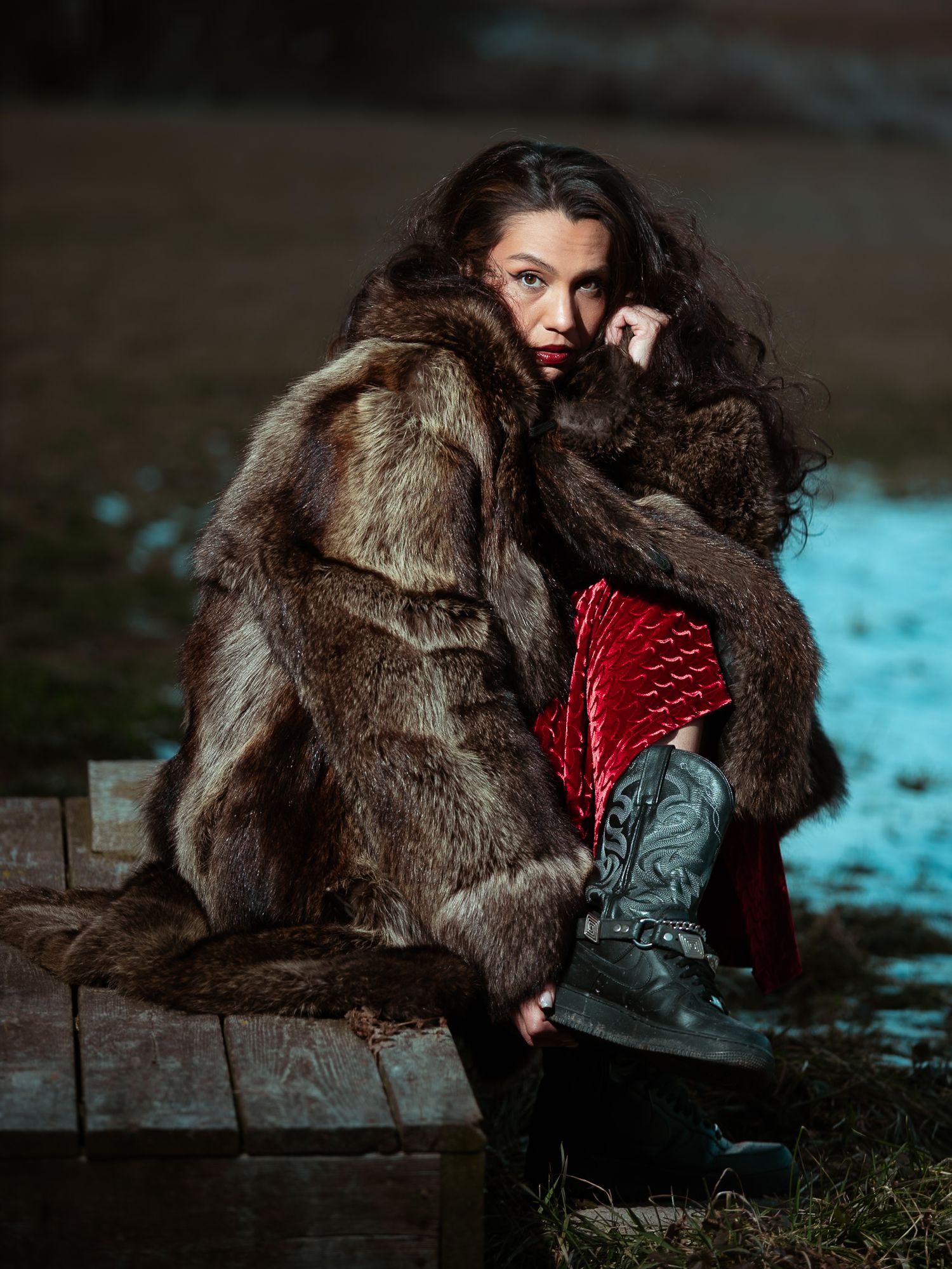 Woman wrapped in oversized brown fur coat wearing red dress and black boots outdoors.