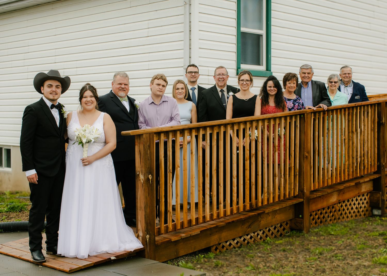 Wedding party poses on wooden deck outside white building with bride in white gown and groom.