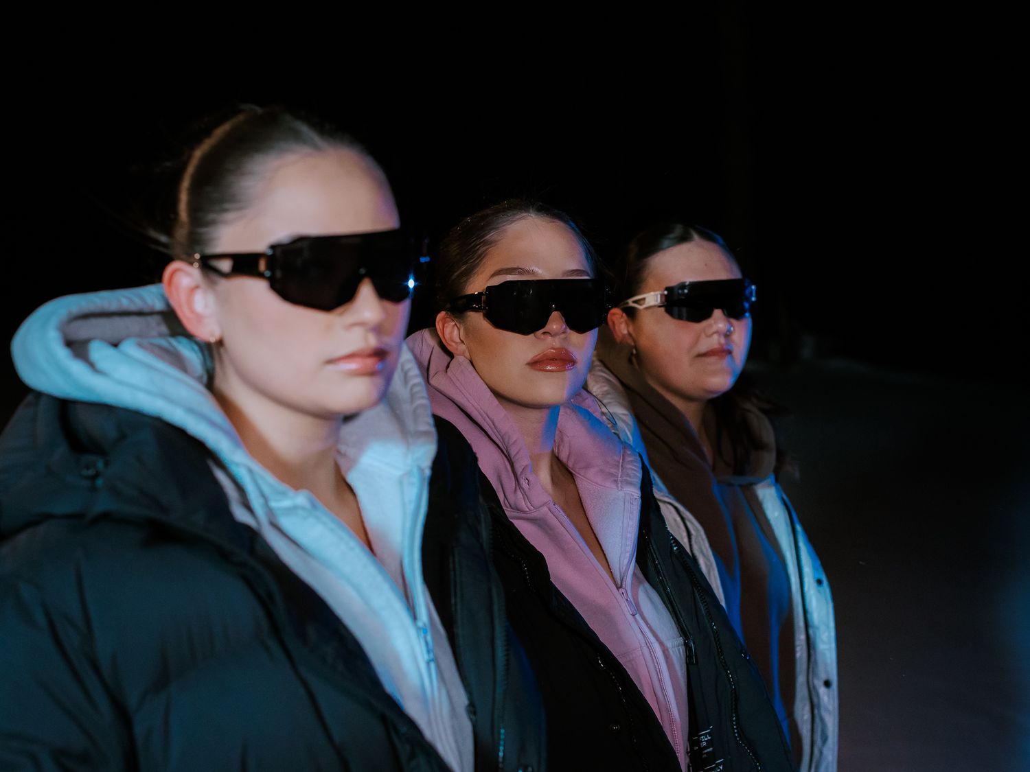Three women wearing black sunglasses and dark puffer jackets in a dramatic nighttime outdoor setting.