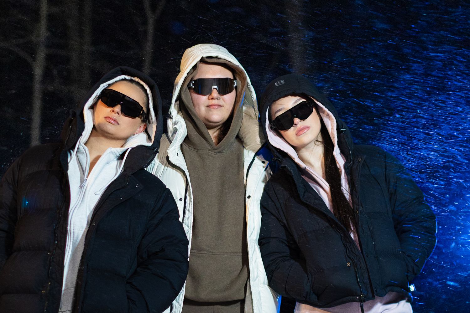 Three people in hooded puffer jackets and black sunglasses posing together in snowy night setting.