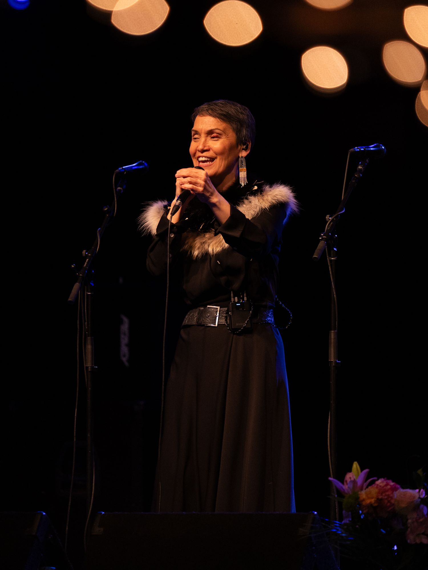 Woman in black outfit with fur trim sings on stage with bokeh lights behind her.