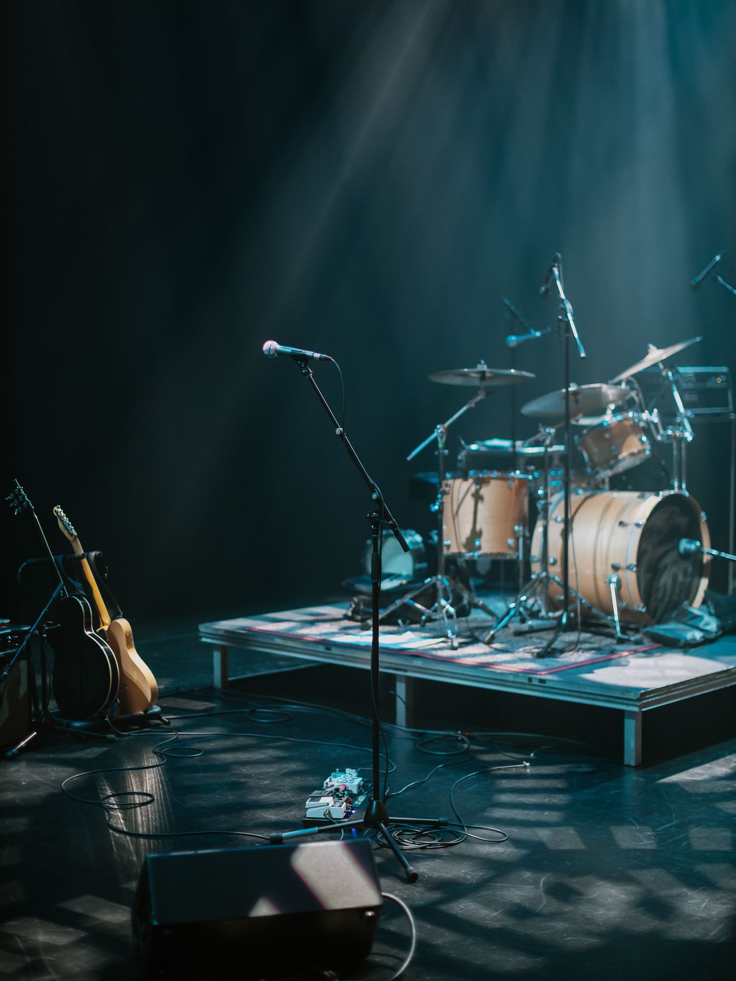 Empty concert stage with drum kit and guitar bathed in dramatic blue spotlights at a performance venue.