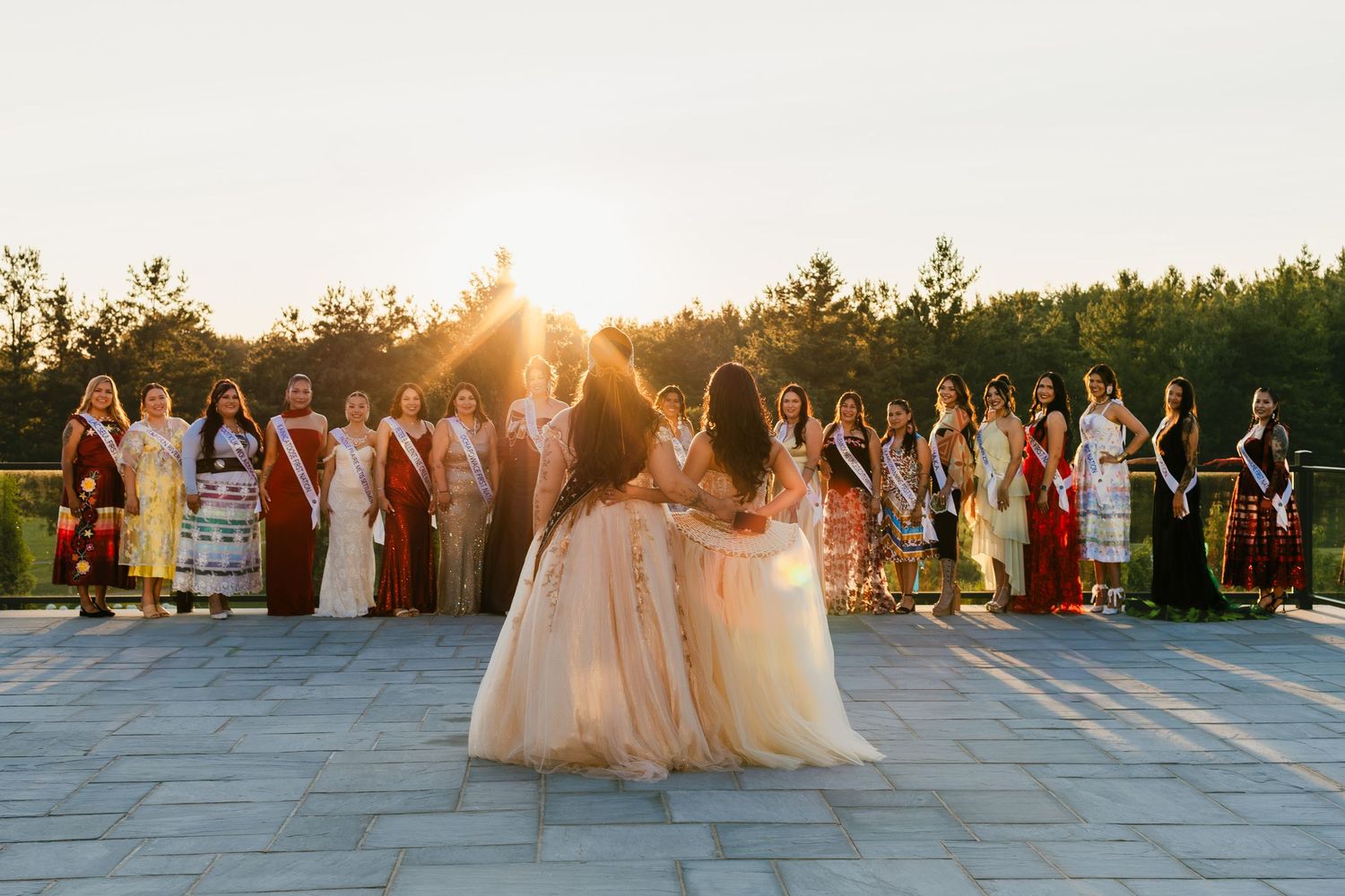 A group of pageant contestants in formal gowns and sashes pose outdoors at golden hour with sunlight streaming through trees.