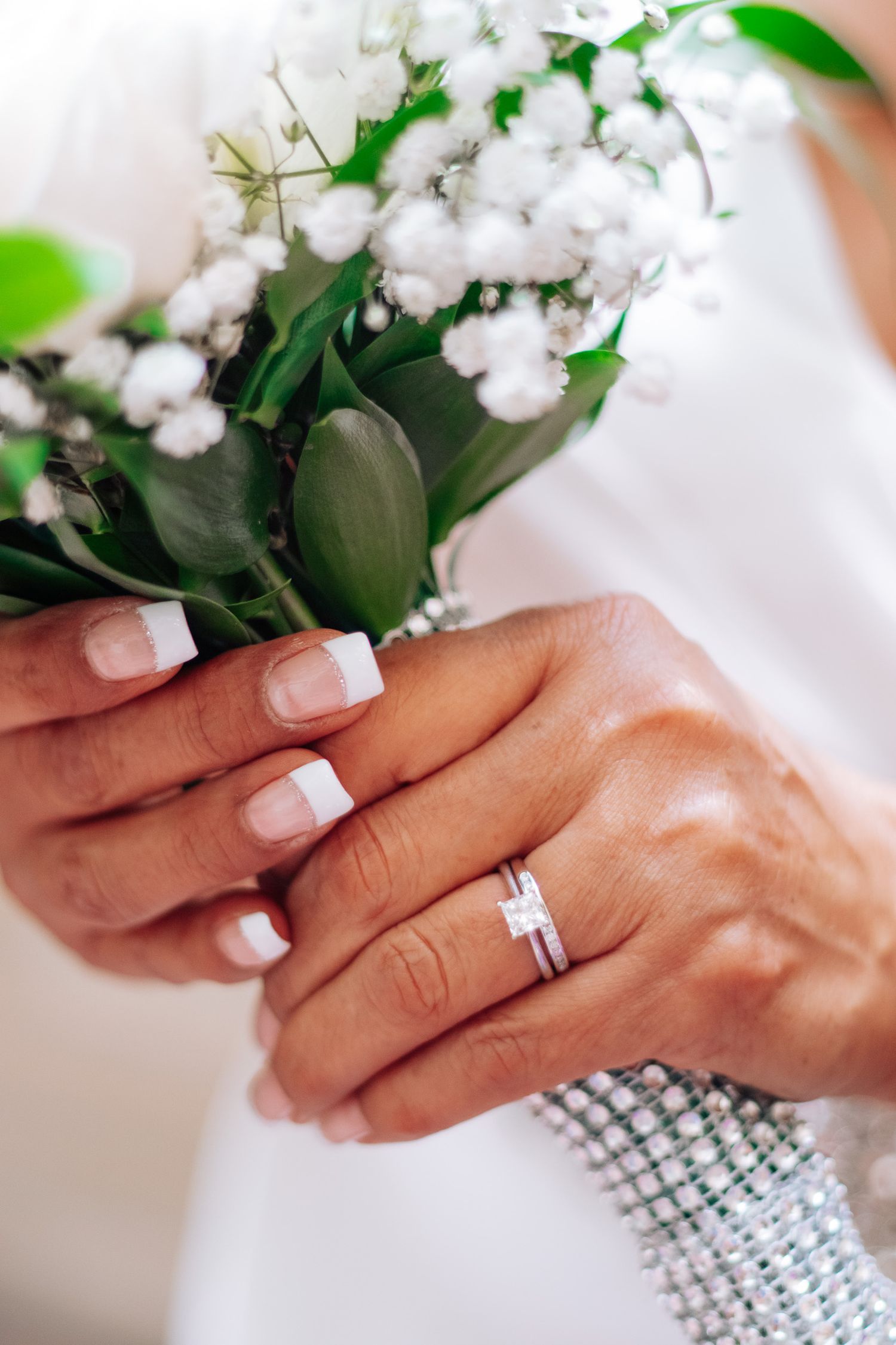 Close-up of bride's hands with French manicure and diamond wedding ring holding white baby's breath bouquet.