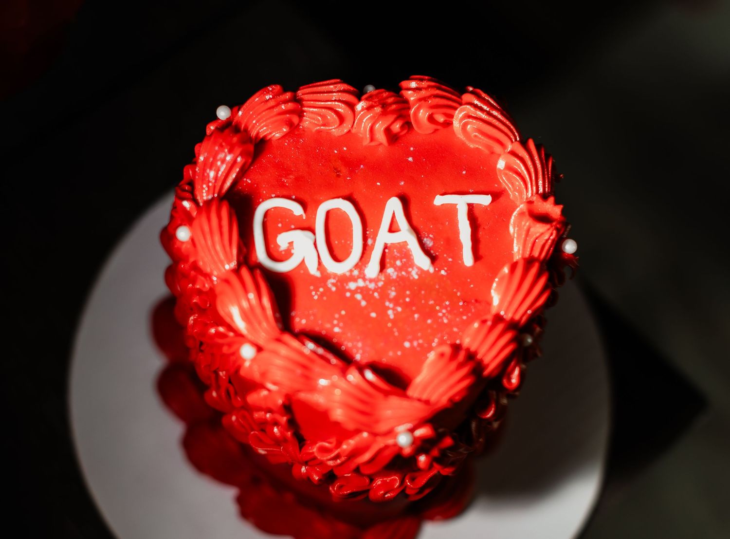 A close-up of a red frosted cake with GOAT text decoration and pearl accents.