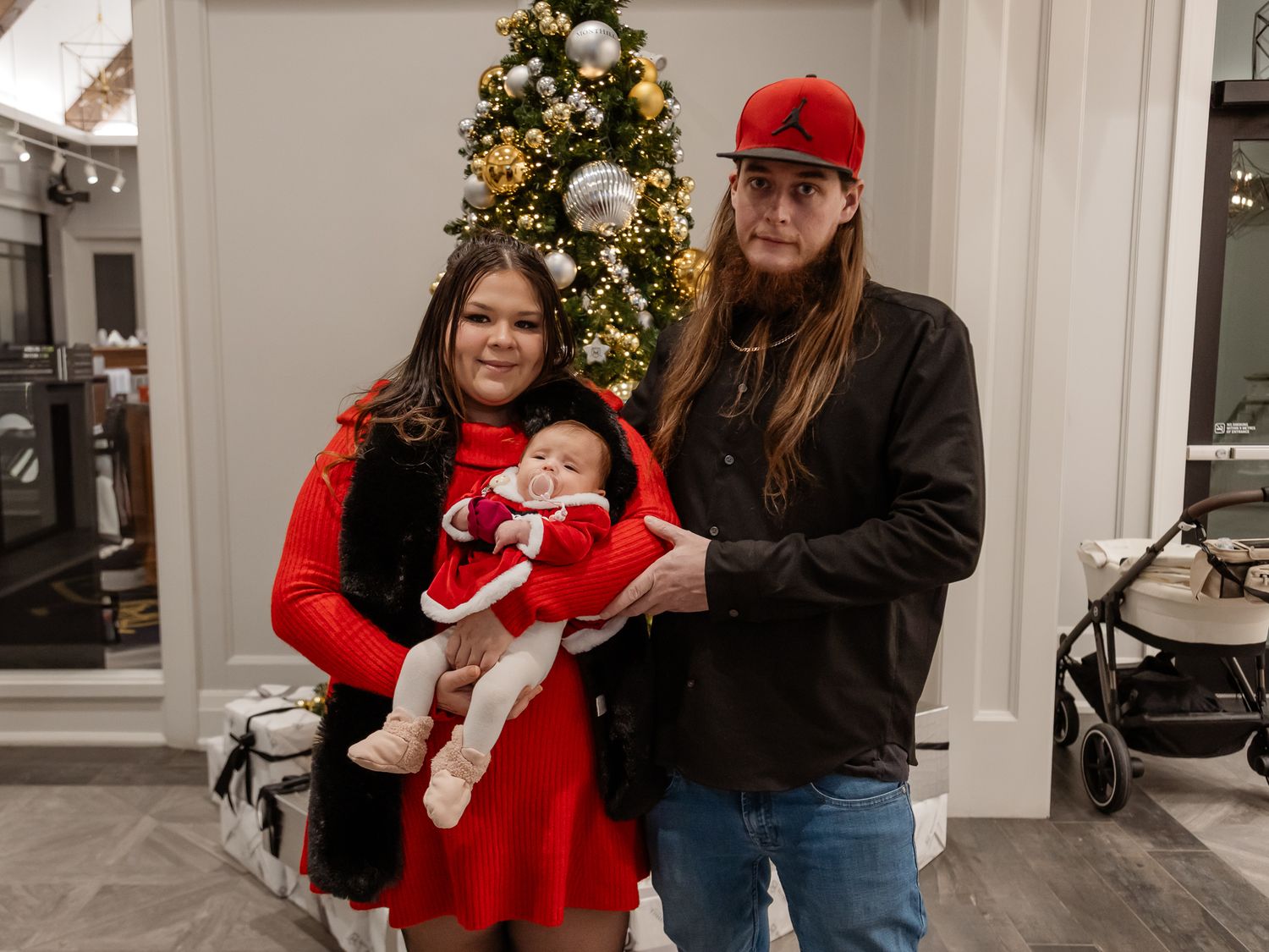 Family poses with baby in holiday attire near decorated Christmas tree at indoor celebration.