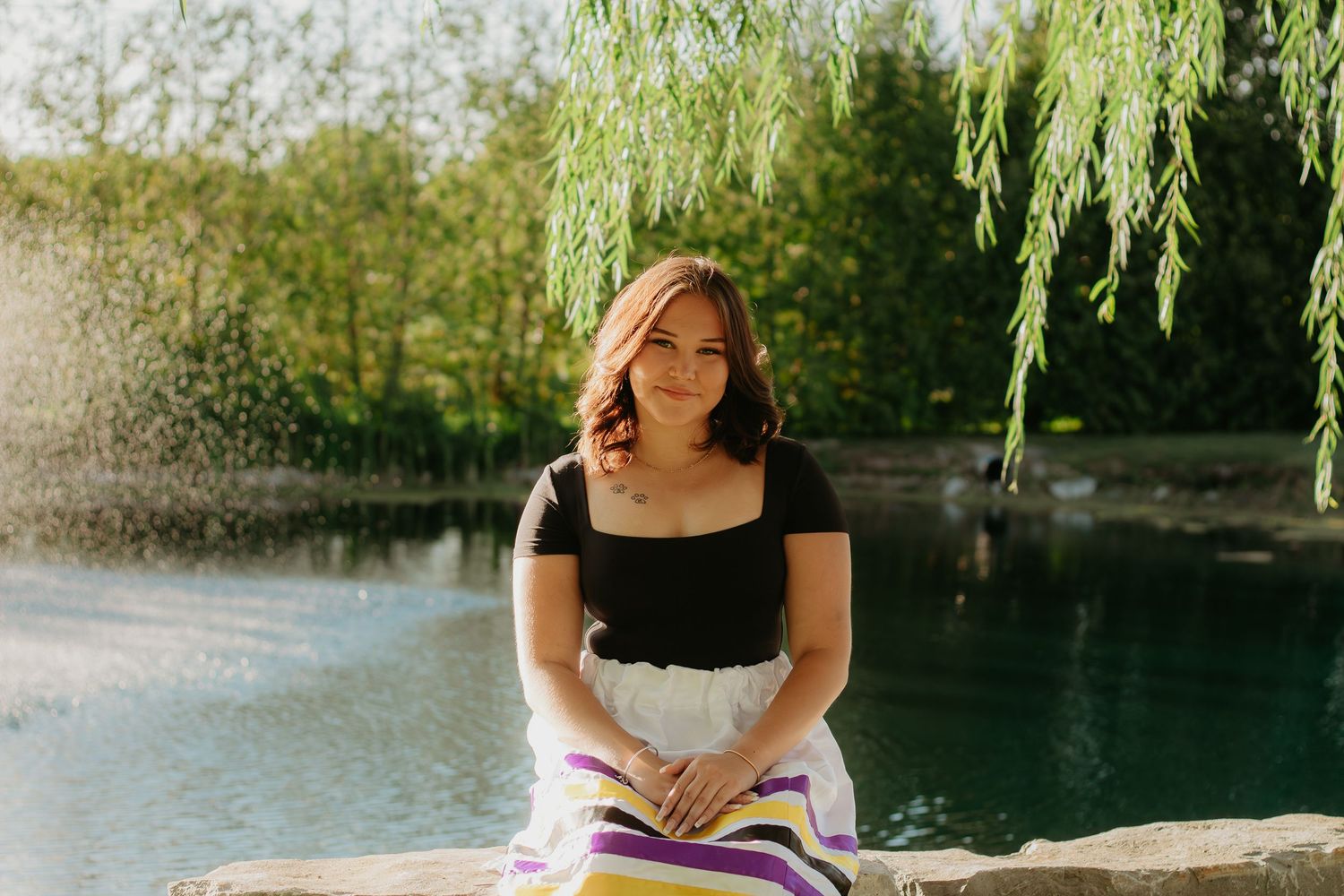Graduate in a black and white dress poses under willow branches near water.
