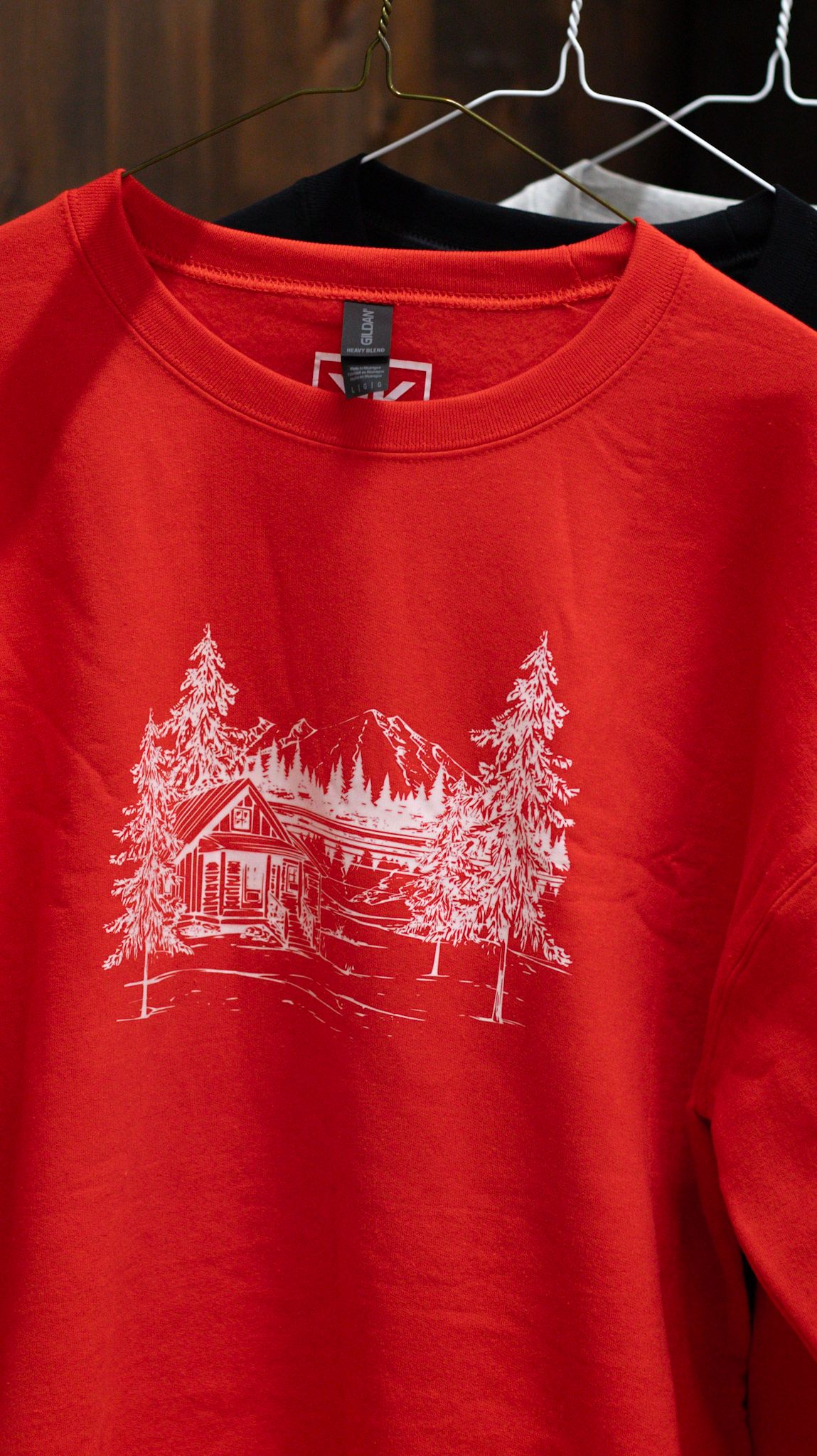 Red sweatshirts featuring white winter cabin scene illustrations hanging on rack.