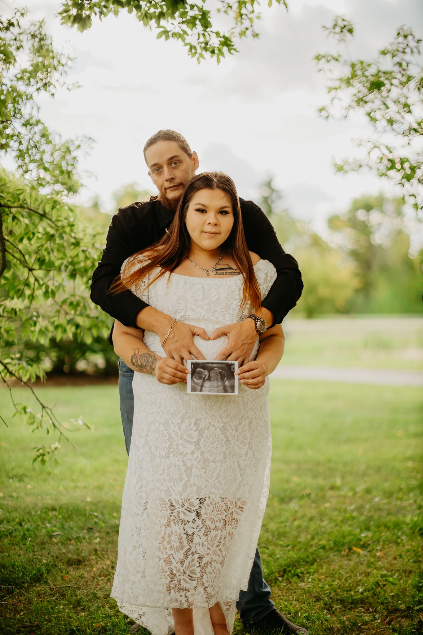 A couple embraces while holding an ultrasound photo in a garden setting.