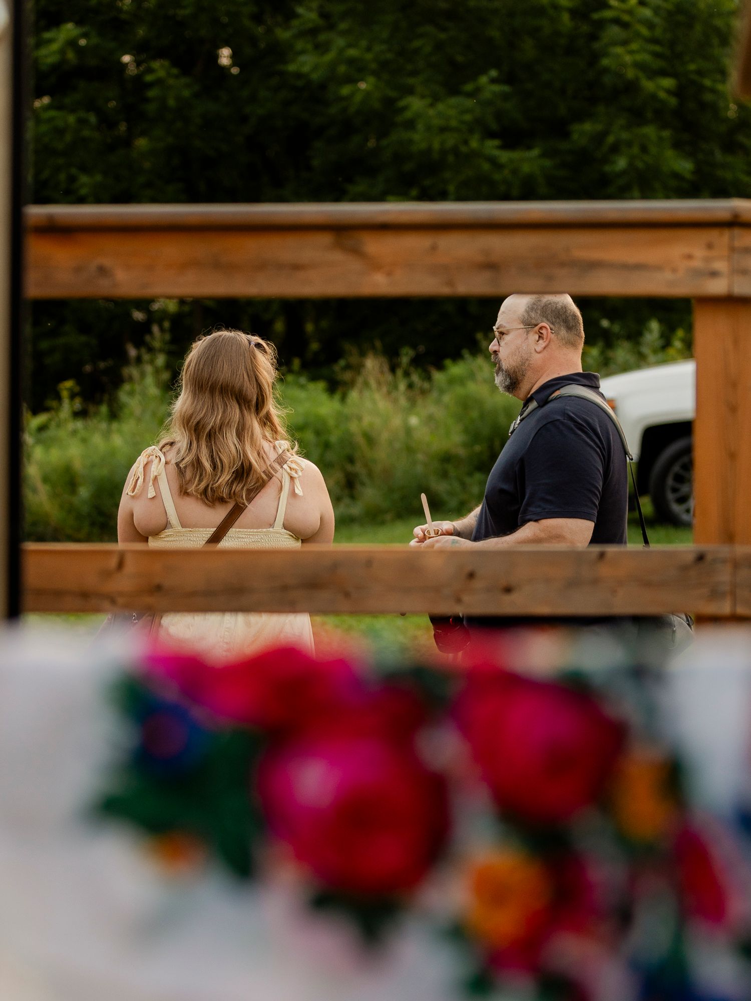 Couple stands before wooden arch at outdoor wedding ceremony with colorful floral arrangements in foreground.