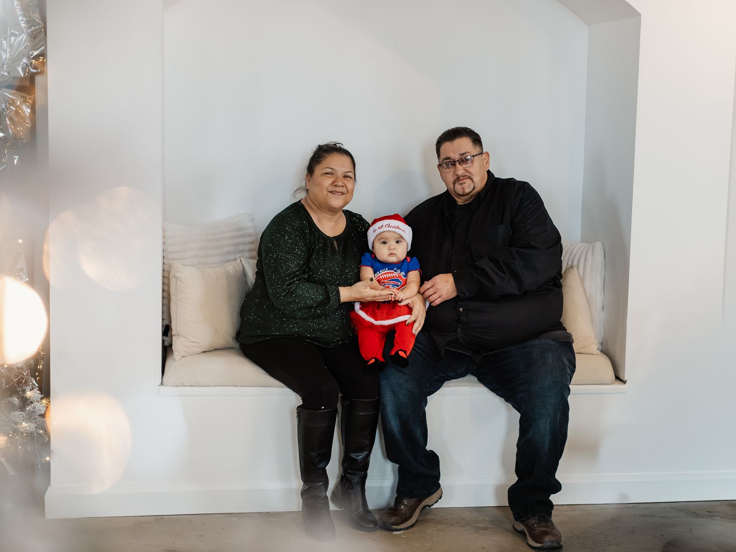 Family of three posing for a Christmas photo, parents holding baby dressed in Santa outfit on a white bench.