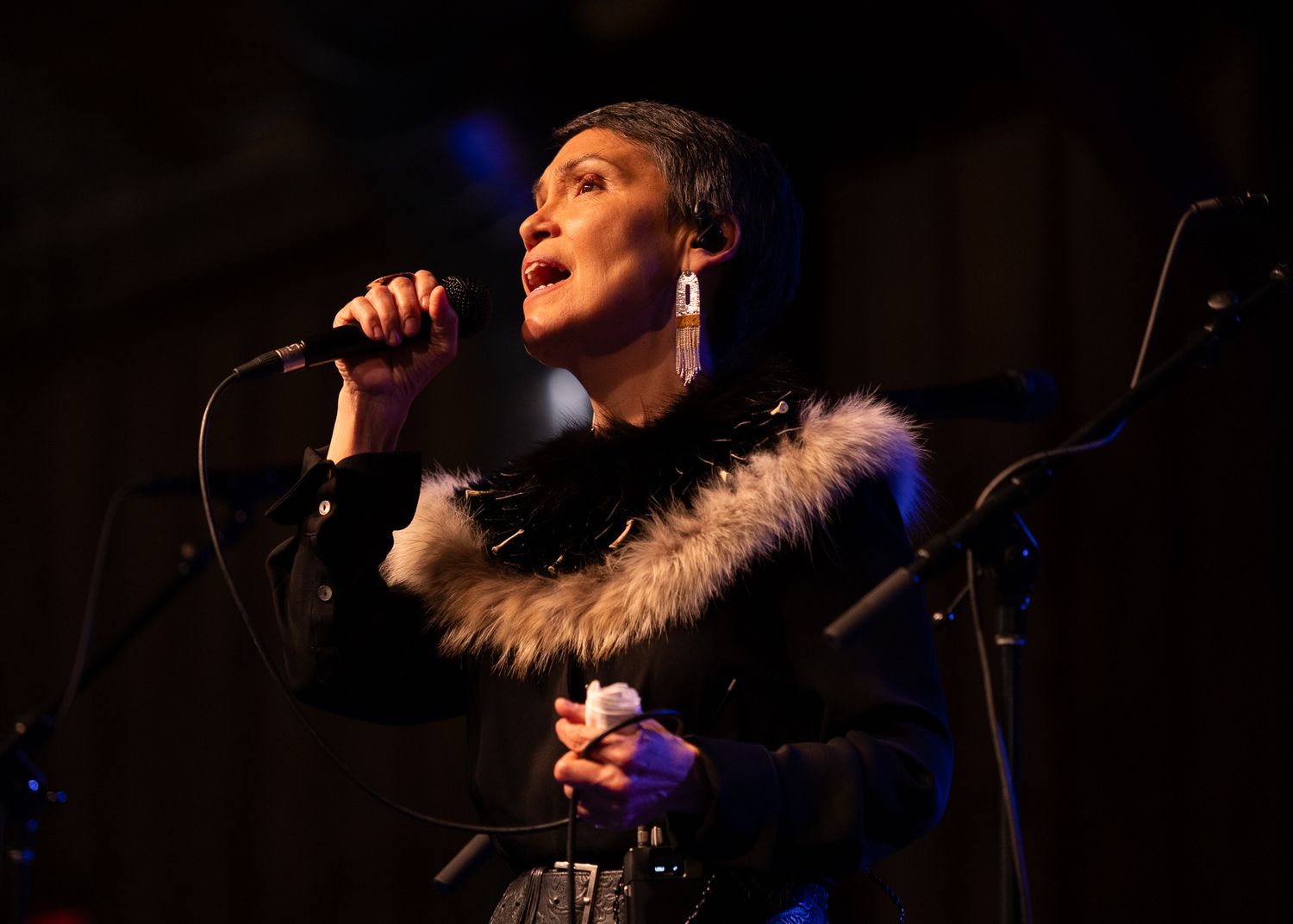 A woman in a black fur-trimmed jacket sings passionately into a microphone on a dimly lit stage.