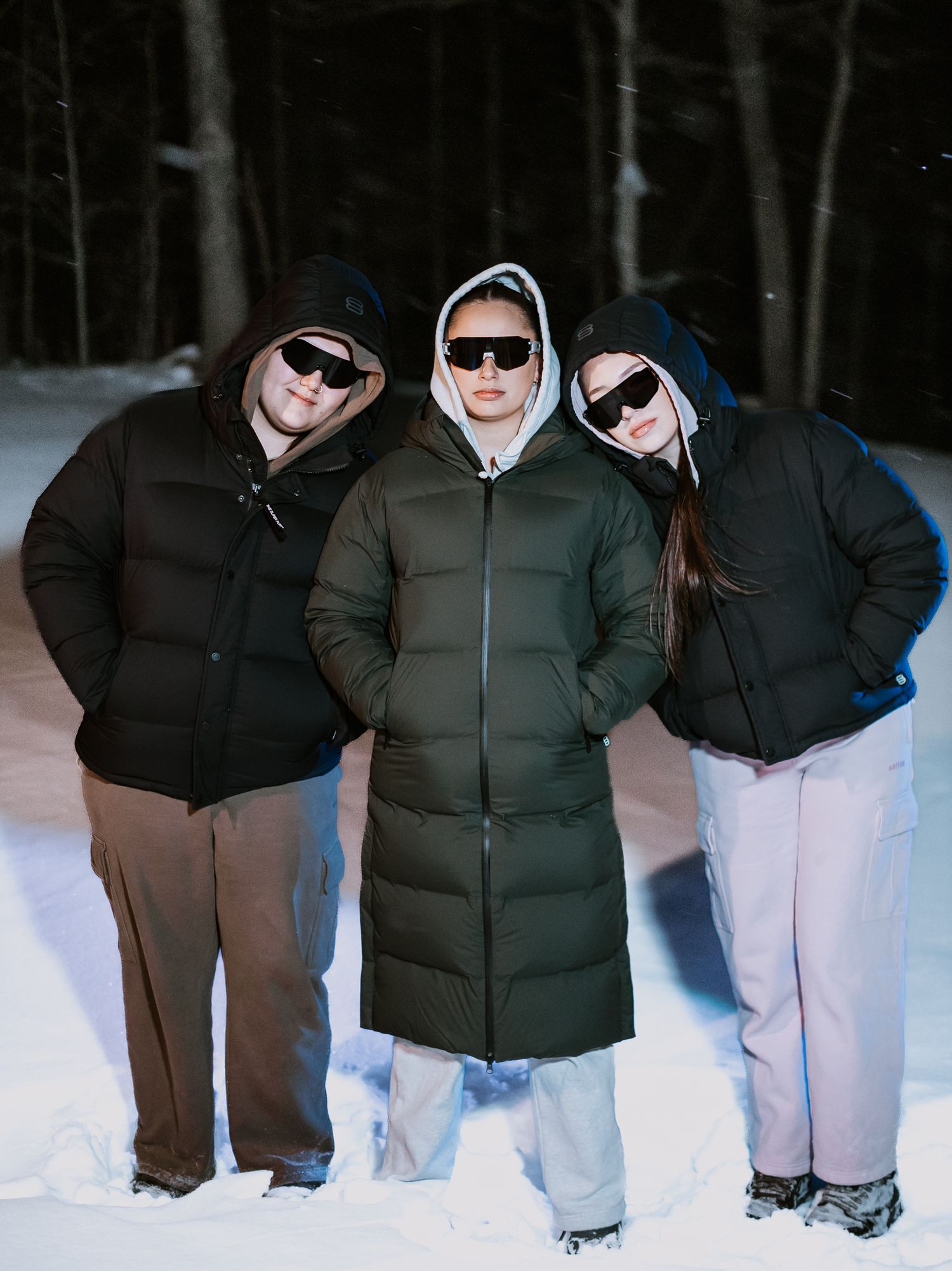 Three models in puffer coats and sunglasses posing together in snowy winter forest.