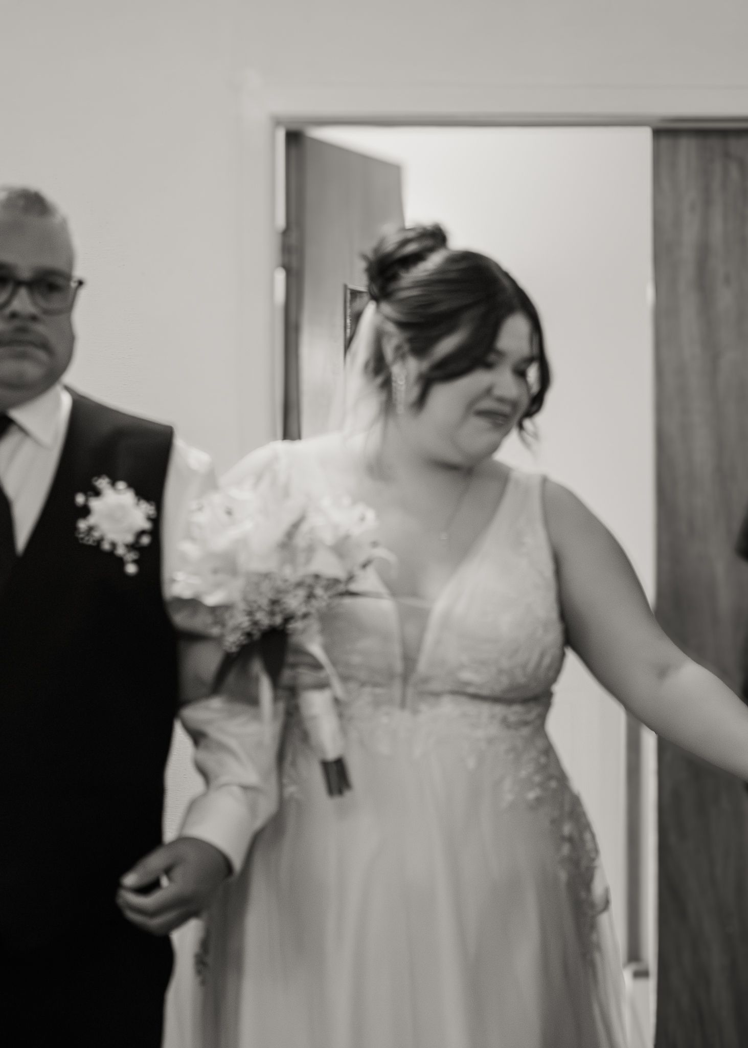 Black and white photo of a bride in a white gown holding a bouquet, walking with a man in a suit.