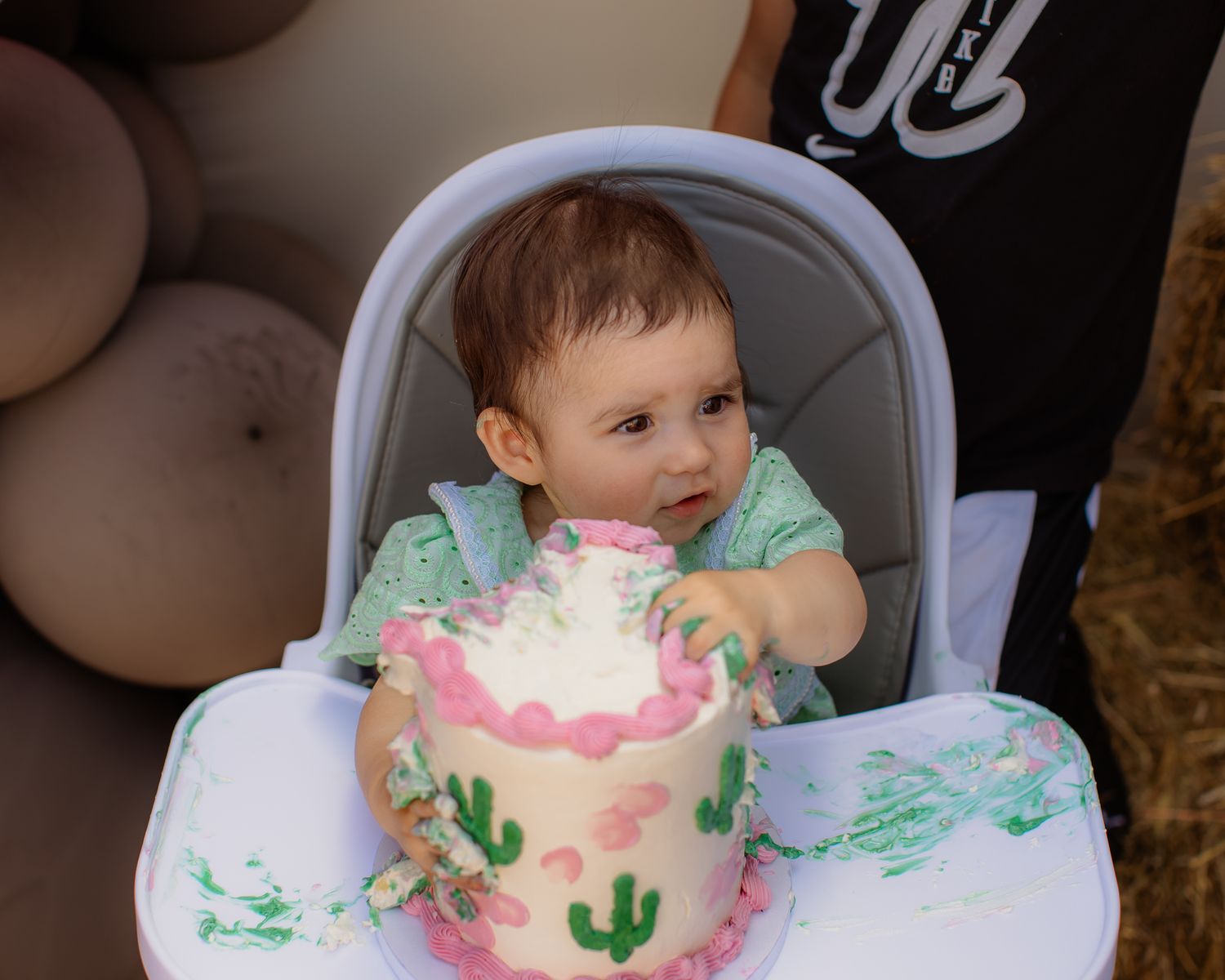 Baby girl in green dress smashes her pink cactus-themed first birthday cake in a white high chair.