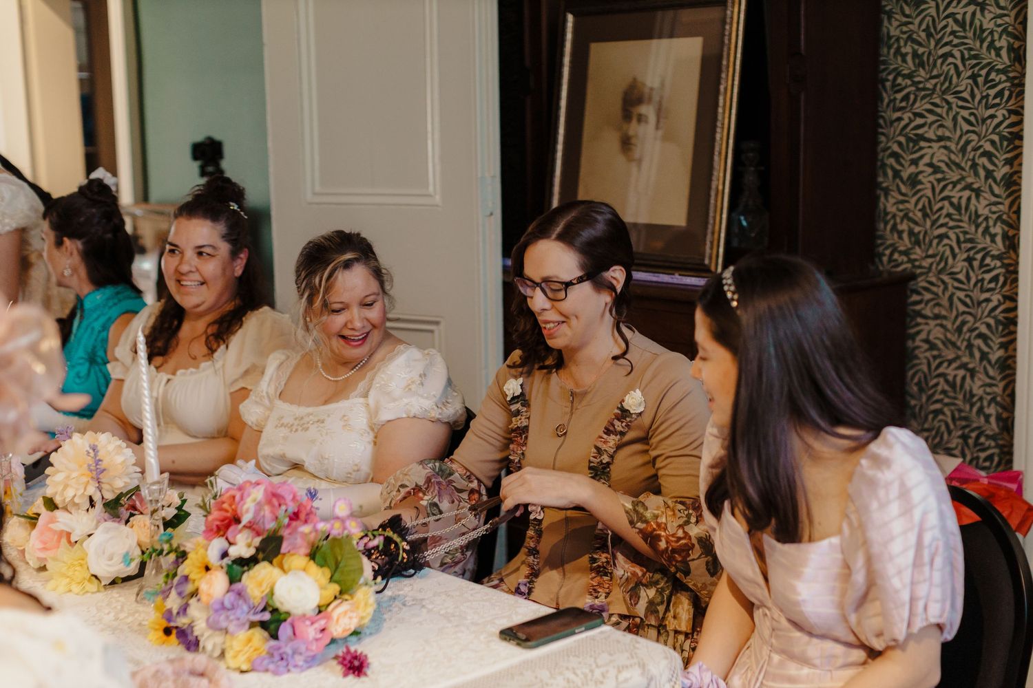 Group of women laughing together at a table with colorful floral arrangements indoors.