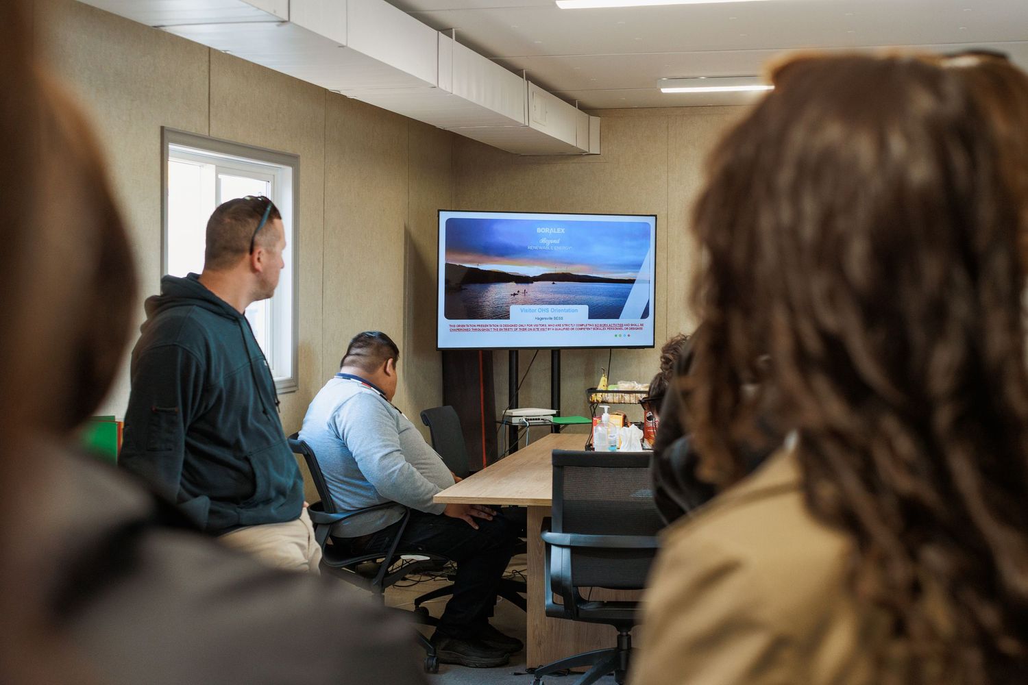 Group of professionals in a conference room watching a presentation on a large wall-mounted screen during a business meeting.