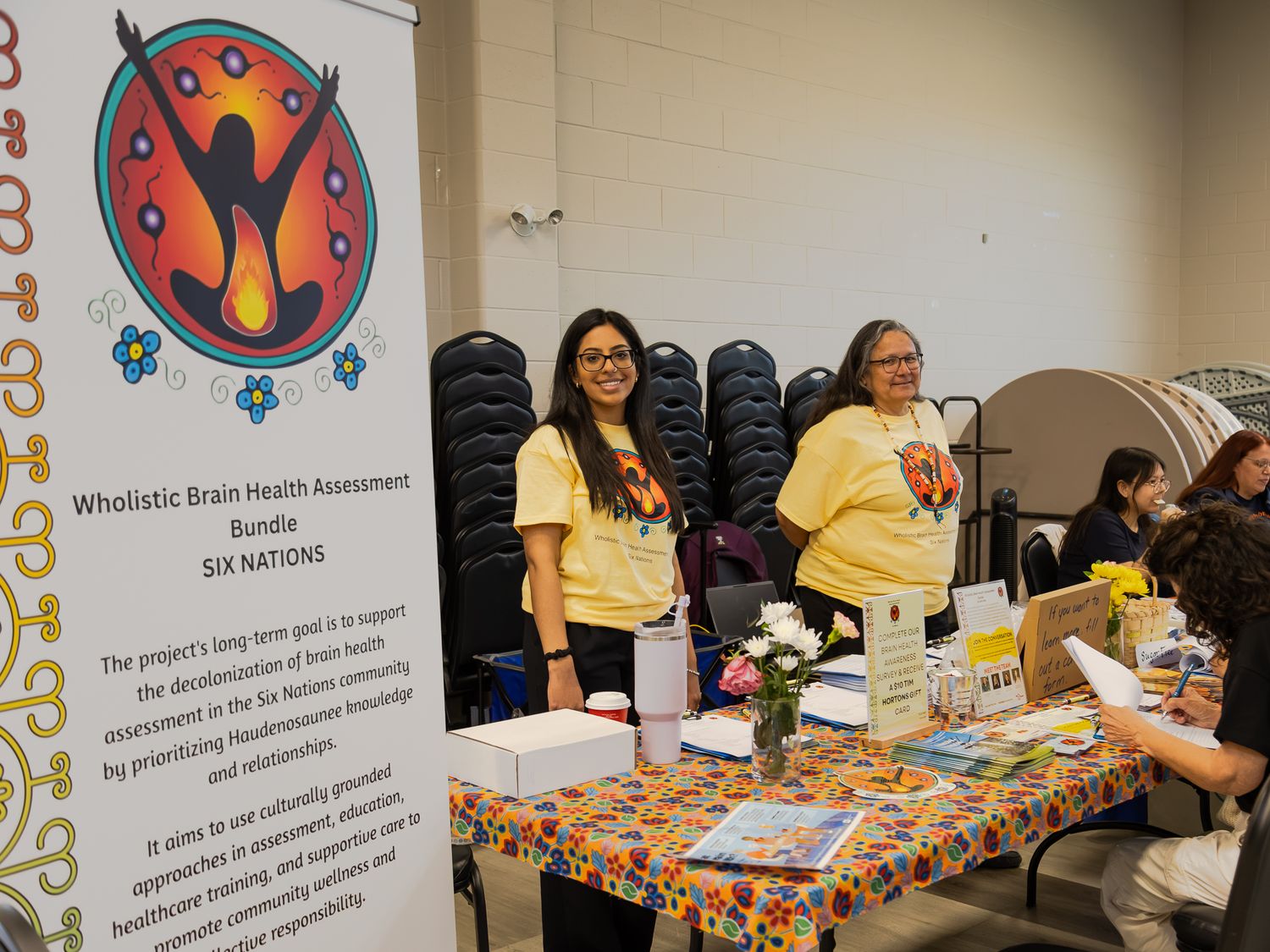 Two women in yellow shirts stand at Six Nations Wholistic Brain Health Assessment Bundle display table.