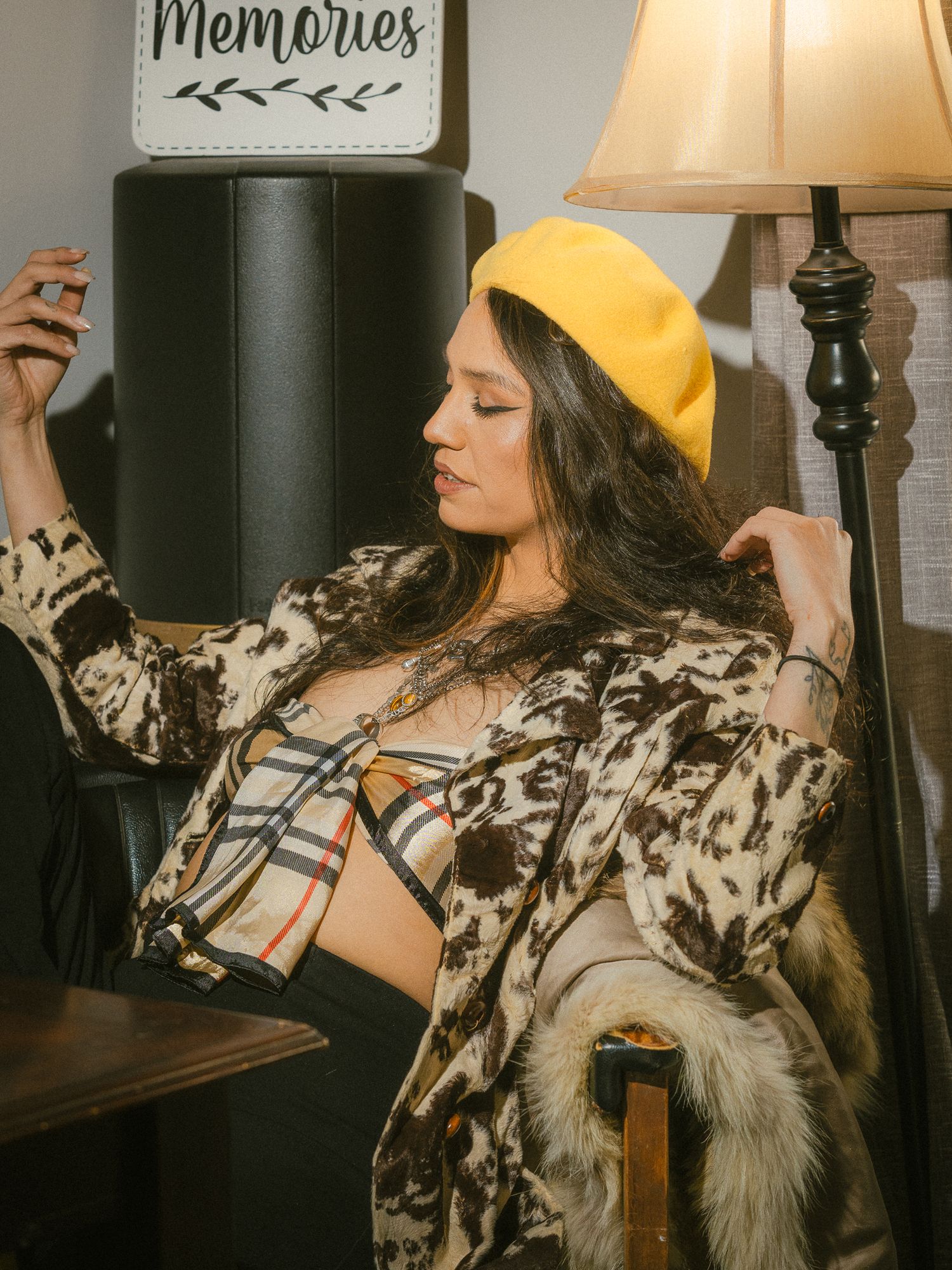 Woman in yellow beret, cow print coat, and Burberry bralette poses indoors near a lamp.