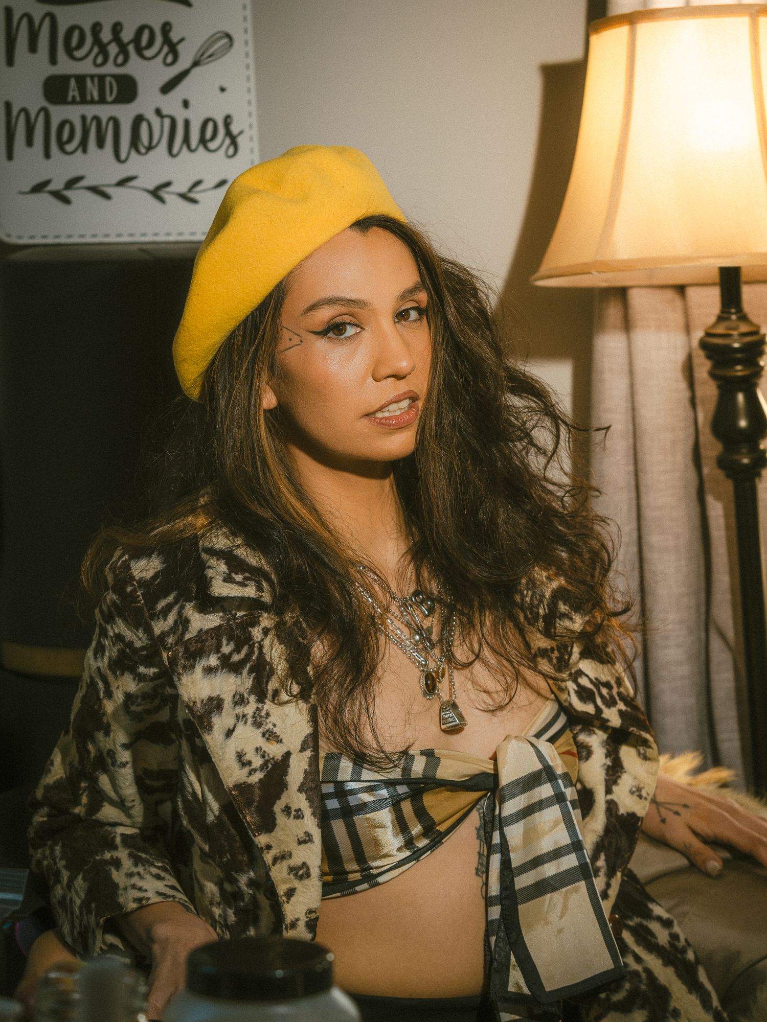 Young woman in yellow beret and plaid jacket sits indoors near lamp, wearing layered necklaces.