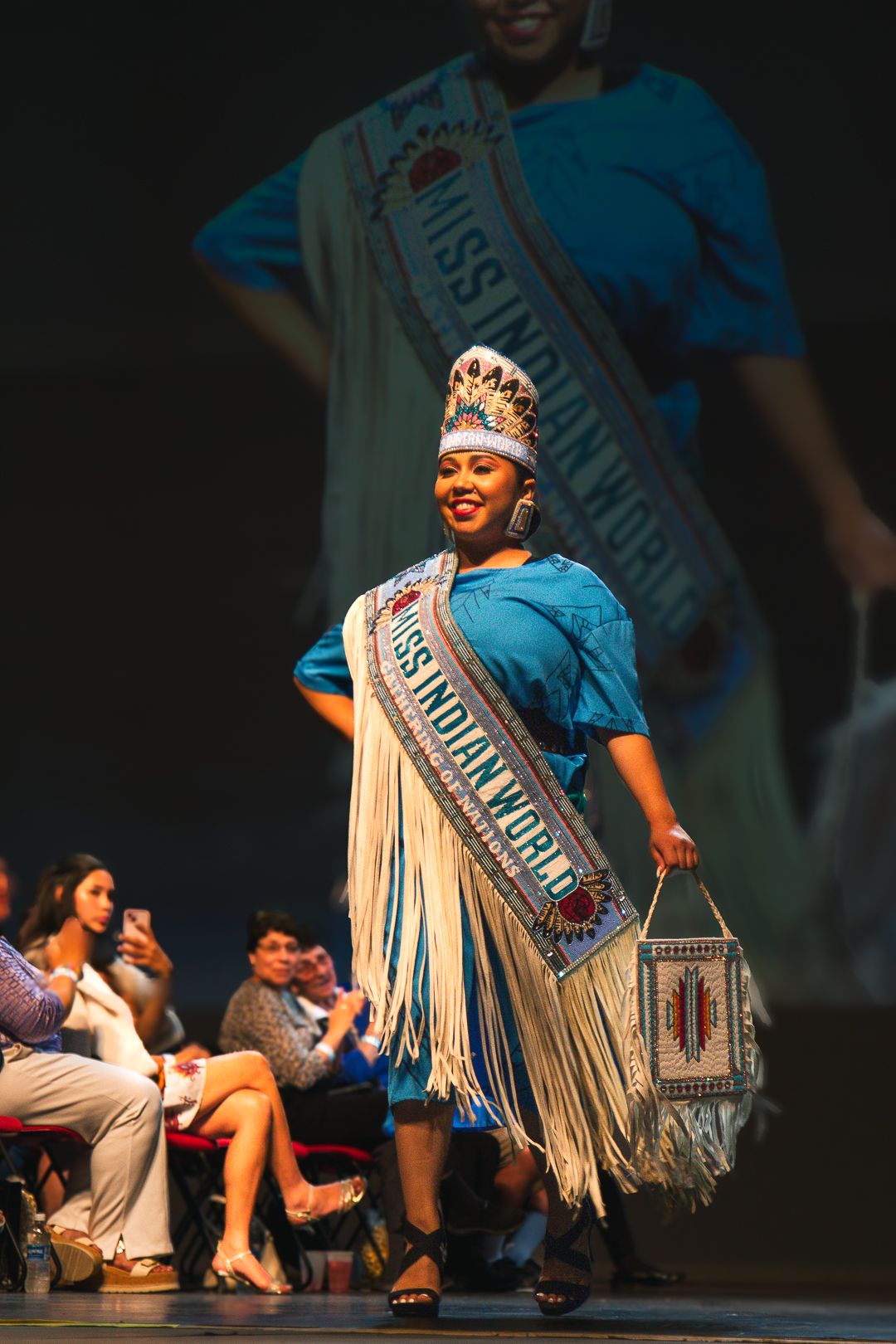 Miss Indian World contestant walks the stage in traditional regalia with fringe sash and beaded crown at pageant.