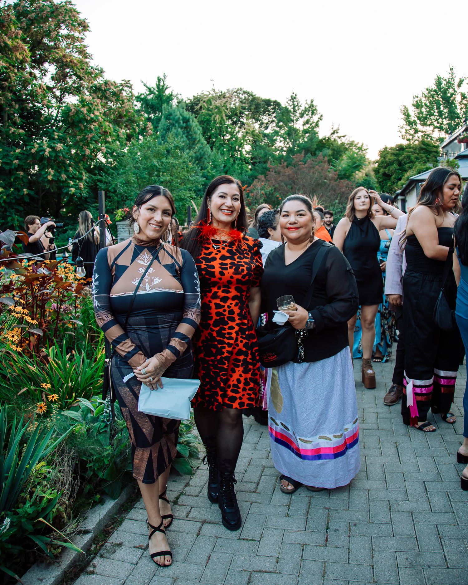 Three women pose outdoors at an evening garden event, dressed in stylish attire including a leopard print dress.