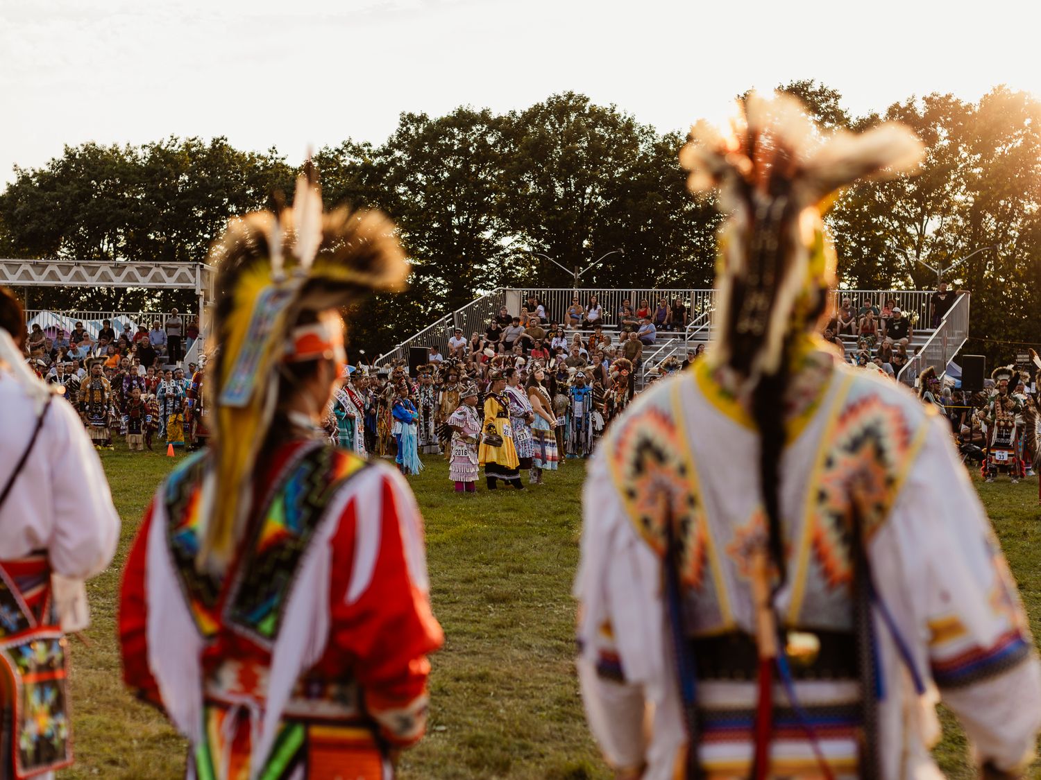Native American dancers in colorful regalia at a sunset powwow with crowd in background.