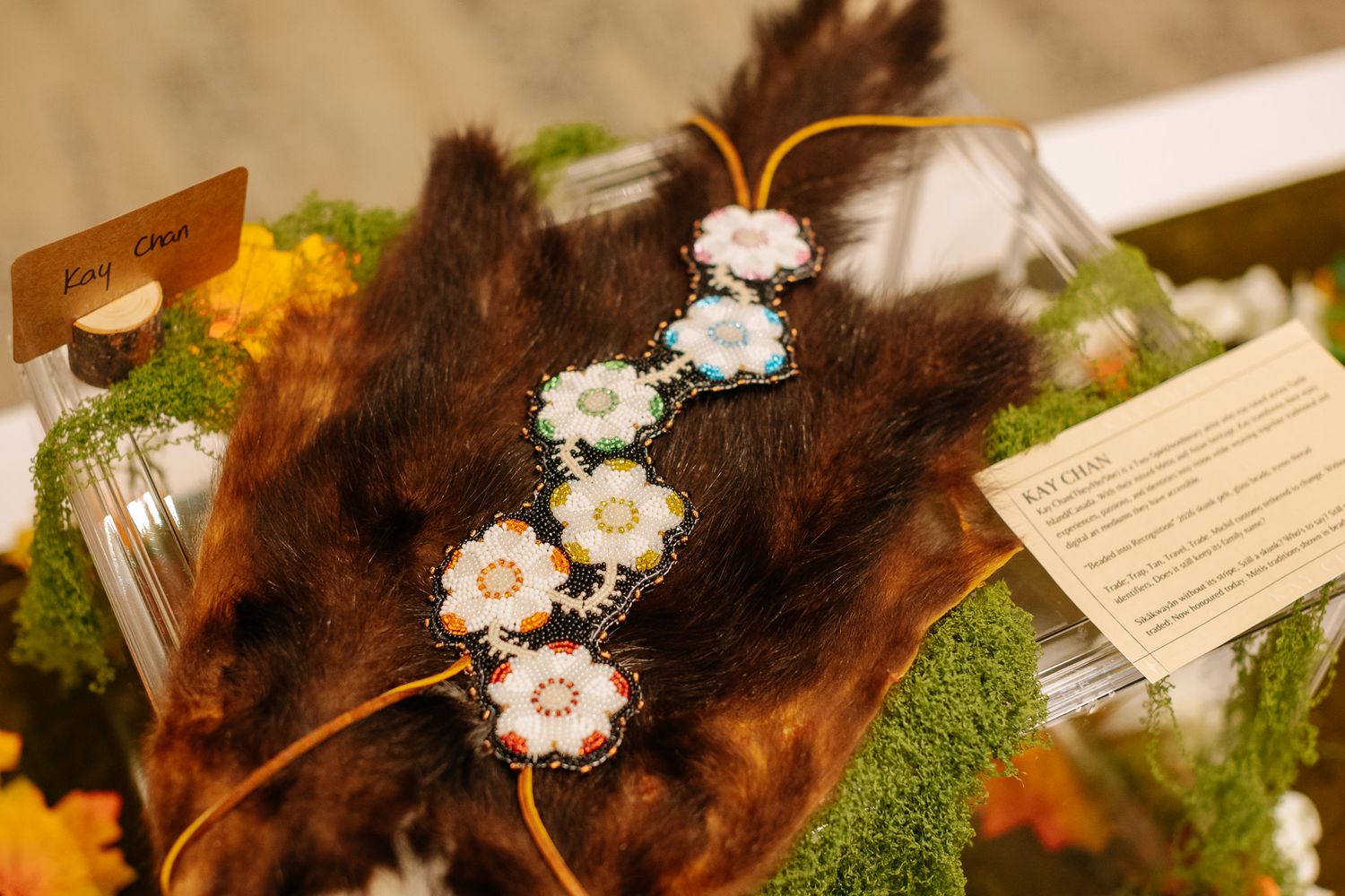 Beaded floral necklace displayed on brown fur with moss and orange flowers at a Native American art exhibit.