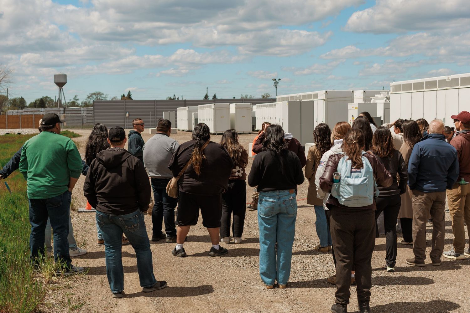 Group of people on a gravel path touring an outdoor industrial facility under a partly cloudy sky.