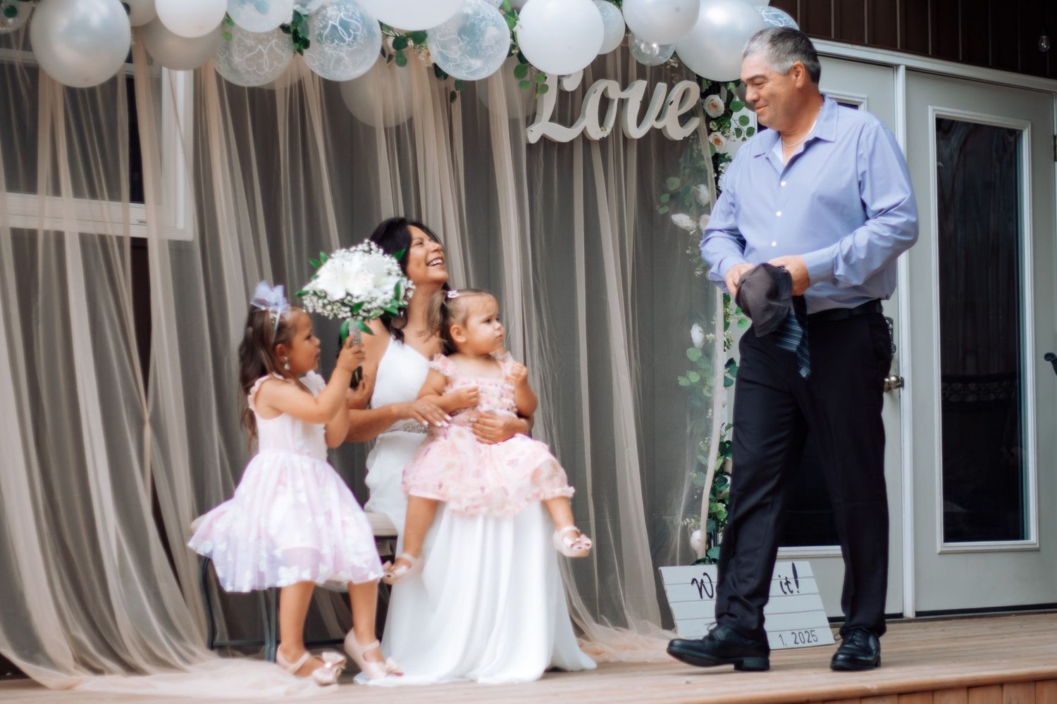 Bride kneeling with two flower girls in pink dresses while man in dark suit looks on at wedding.