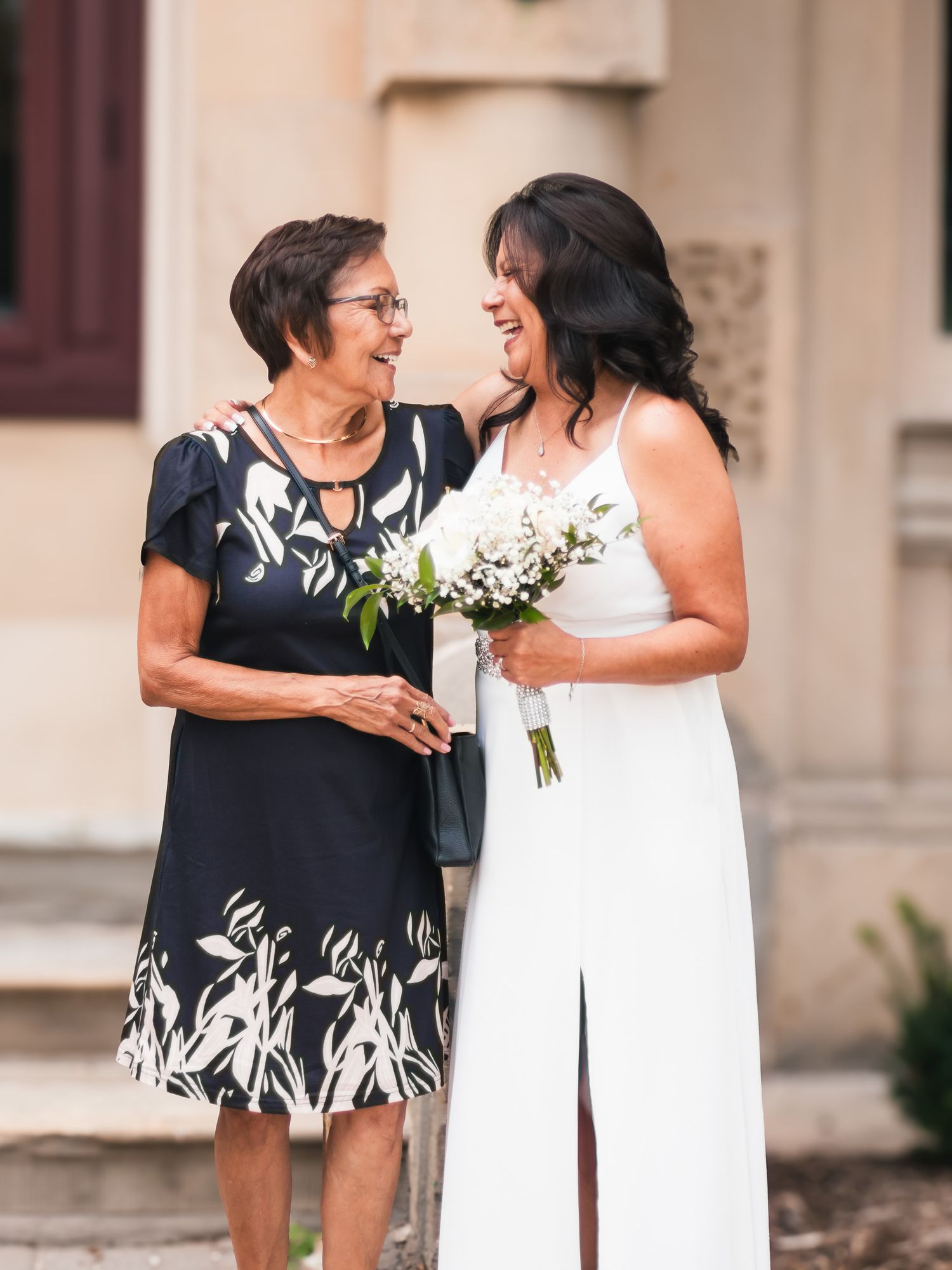 Bride in white gown laughing with older woman in navy floral dress, sharing a joyful moment outdoors.