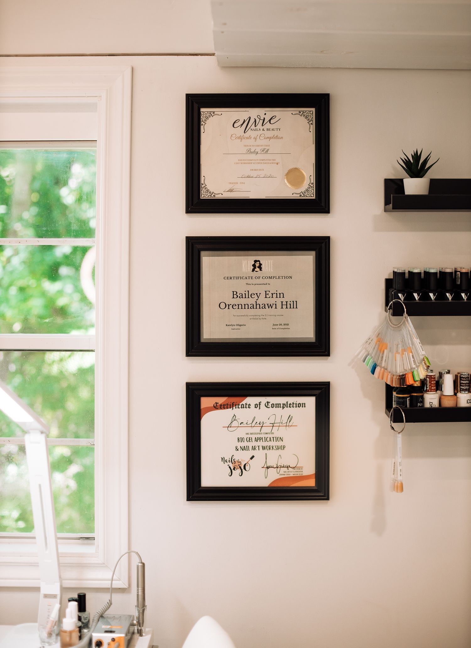 Three framed certificates hang on a white wall next to a window with decorative shelving.