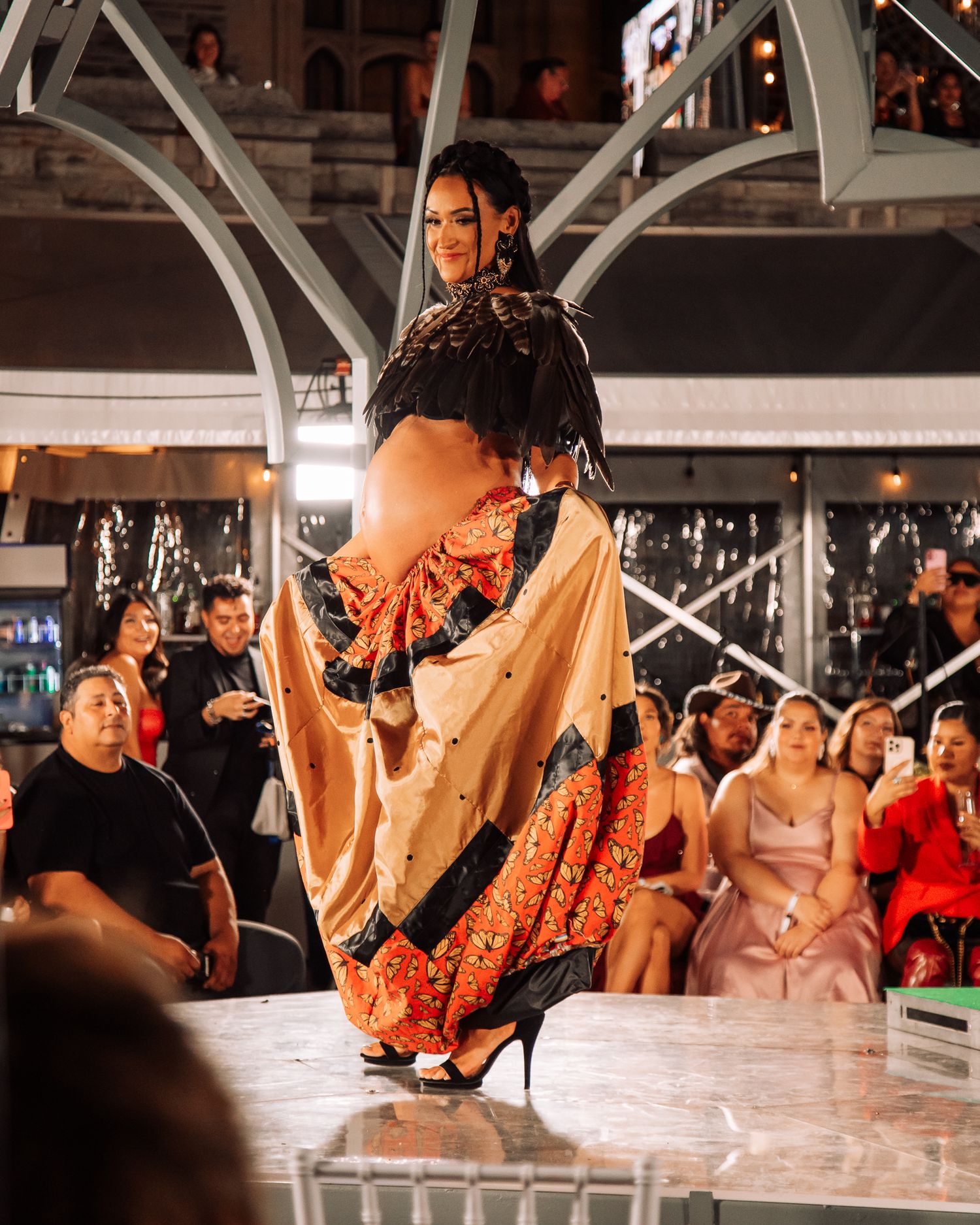 Fashion models display elegant orange and black evening wear on a reflective runway surface.