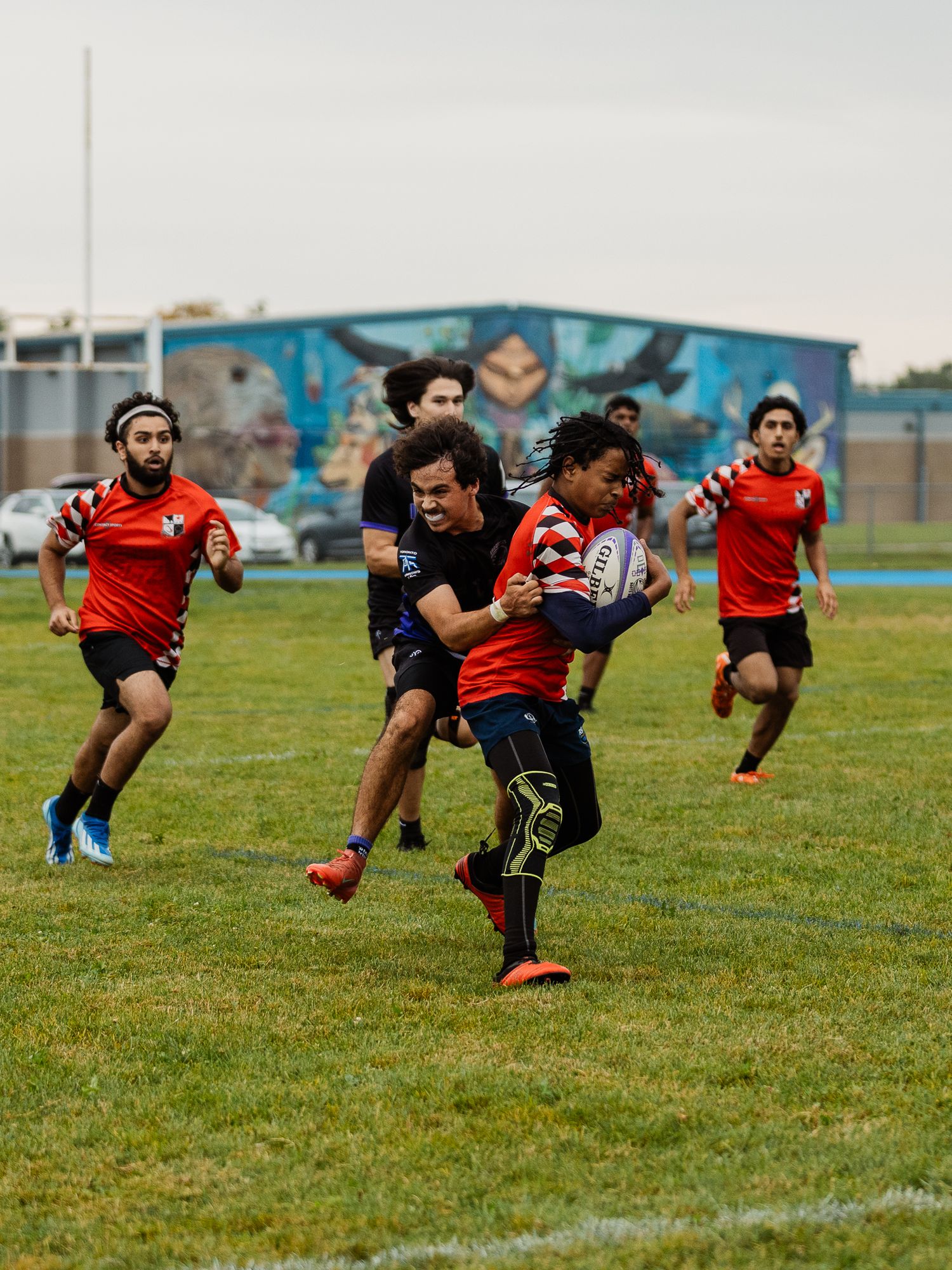 Rugby player in red jersey carries ball while being tackled during an outdoor match with teammates running in support.