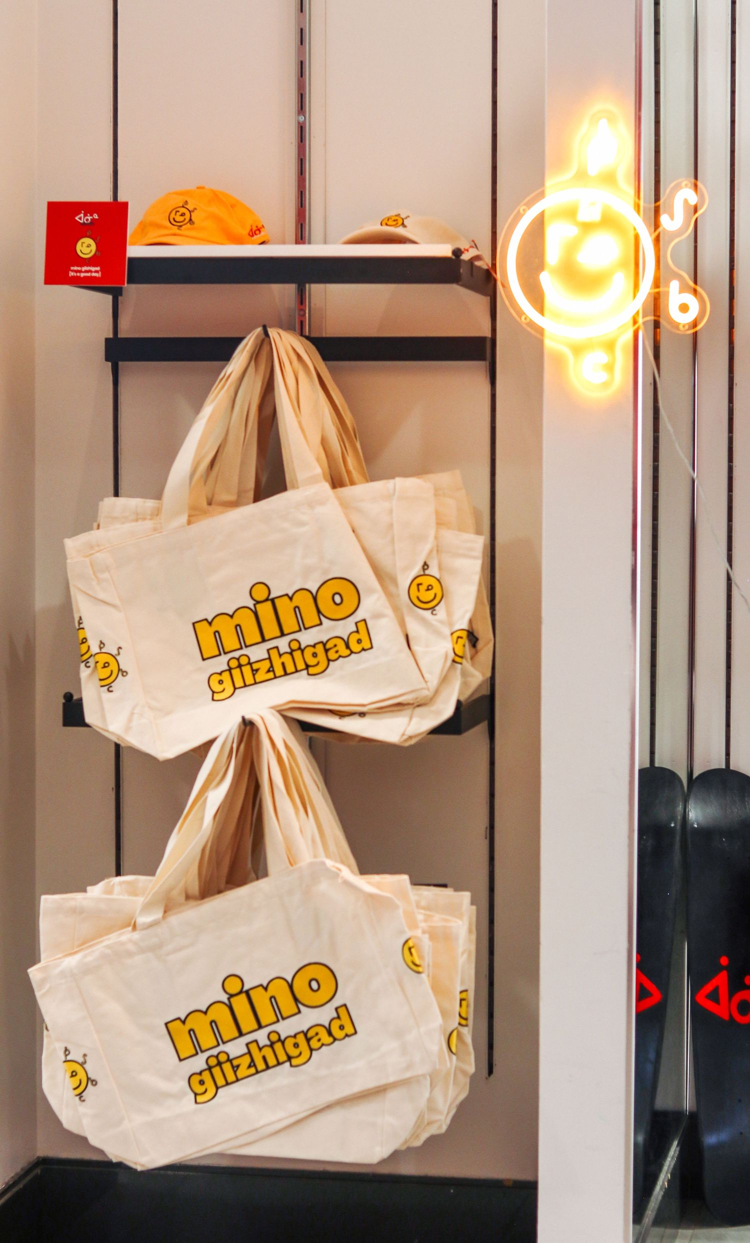 Canvas tote bags with yellow 'mino giizhigad' text hang on a rack beside a glowing neon sign in a retail store.