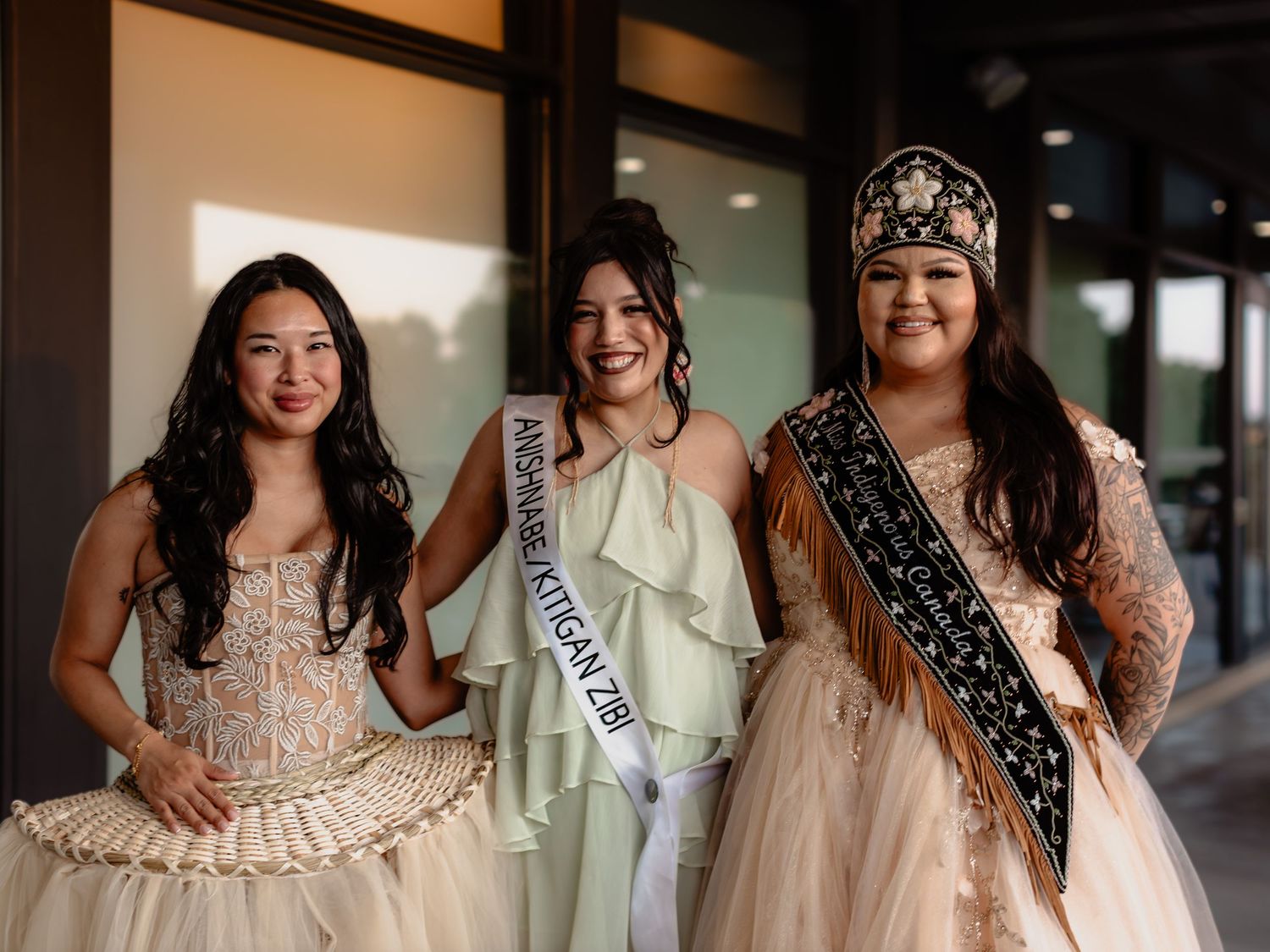 Three women in formal gowns, two wearing pageant sashes, posing together outdoors at a beauty competition.
