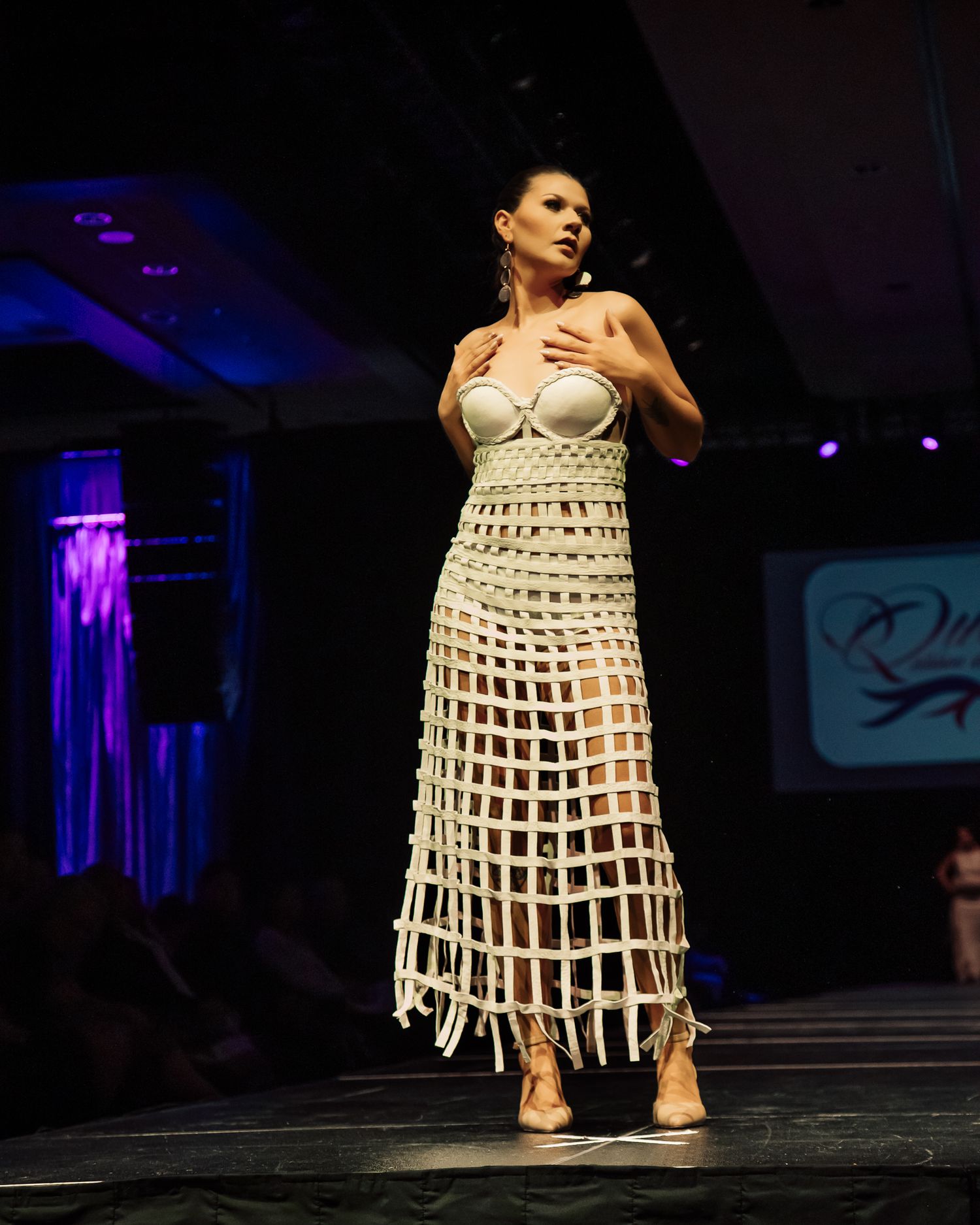 Model walks runway in avant-garde white grid-pattern dress at fashion show event.