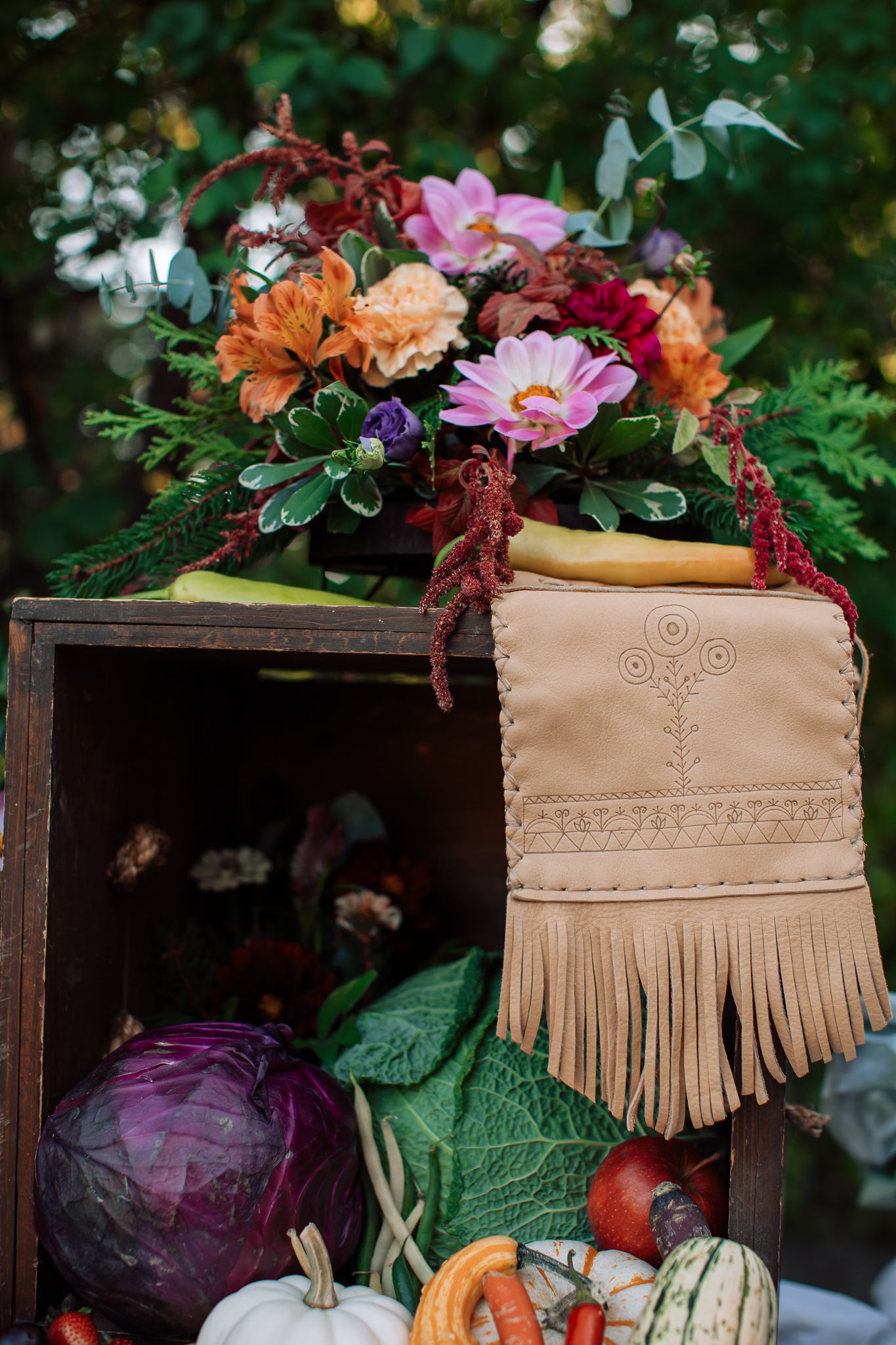Decorative display of colorful flowers, vegetables, and fringed leather bag in rustic wooden crate.