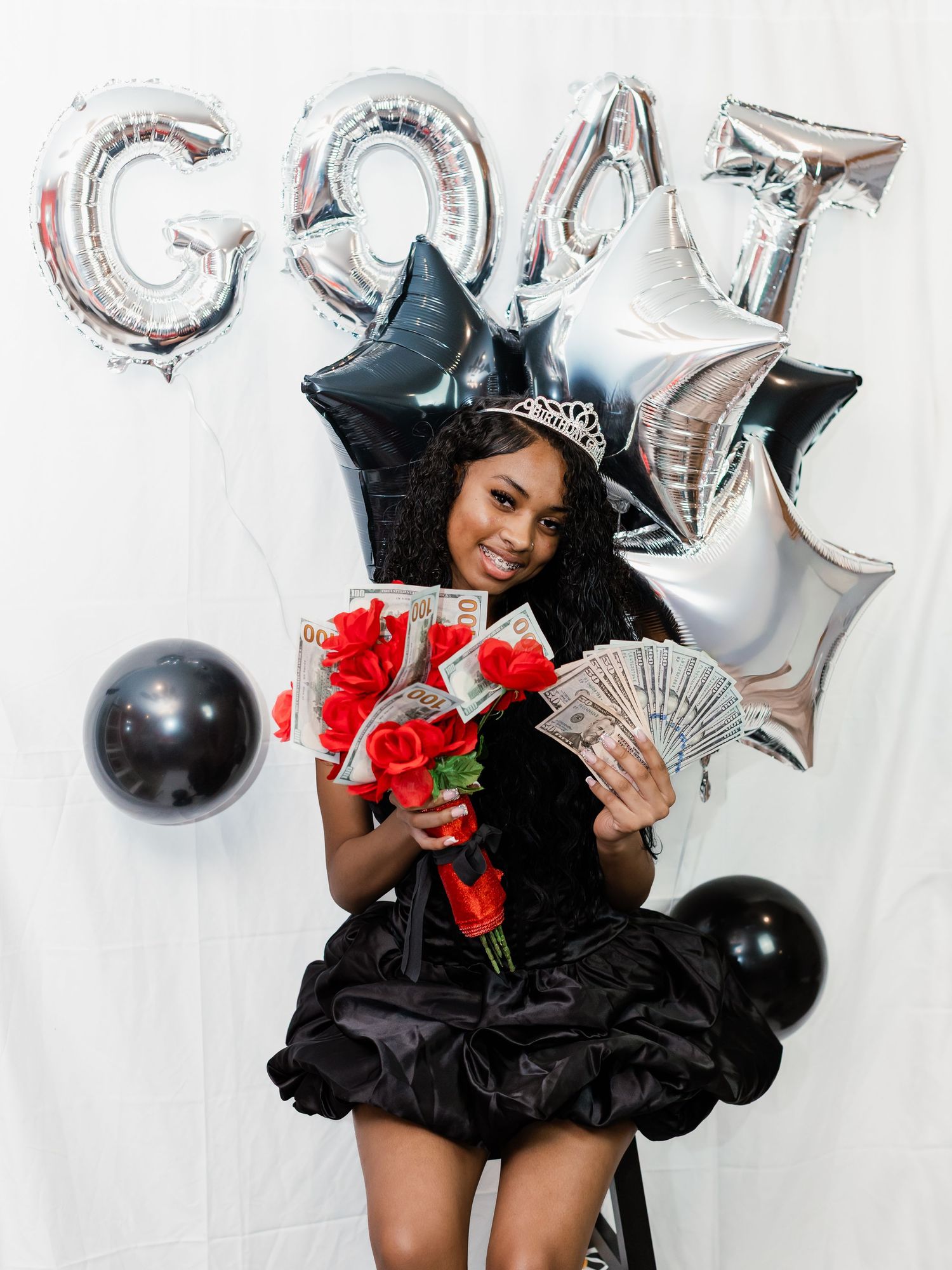 Young woman in black dress and tiara holds red roses and cash in front of GOAT balloons at a celebration.
