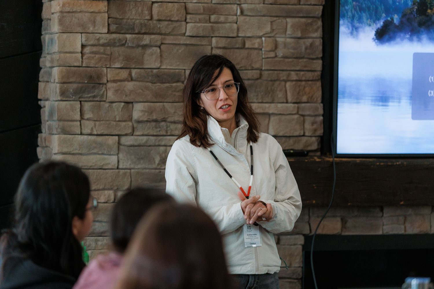 Woman in white jacket presenting at a conference near a brick wall with a projected screen behind her.