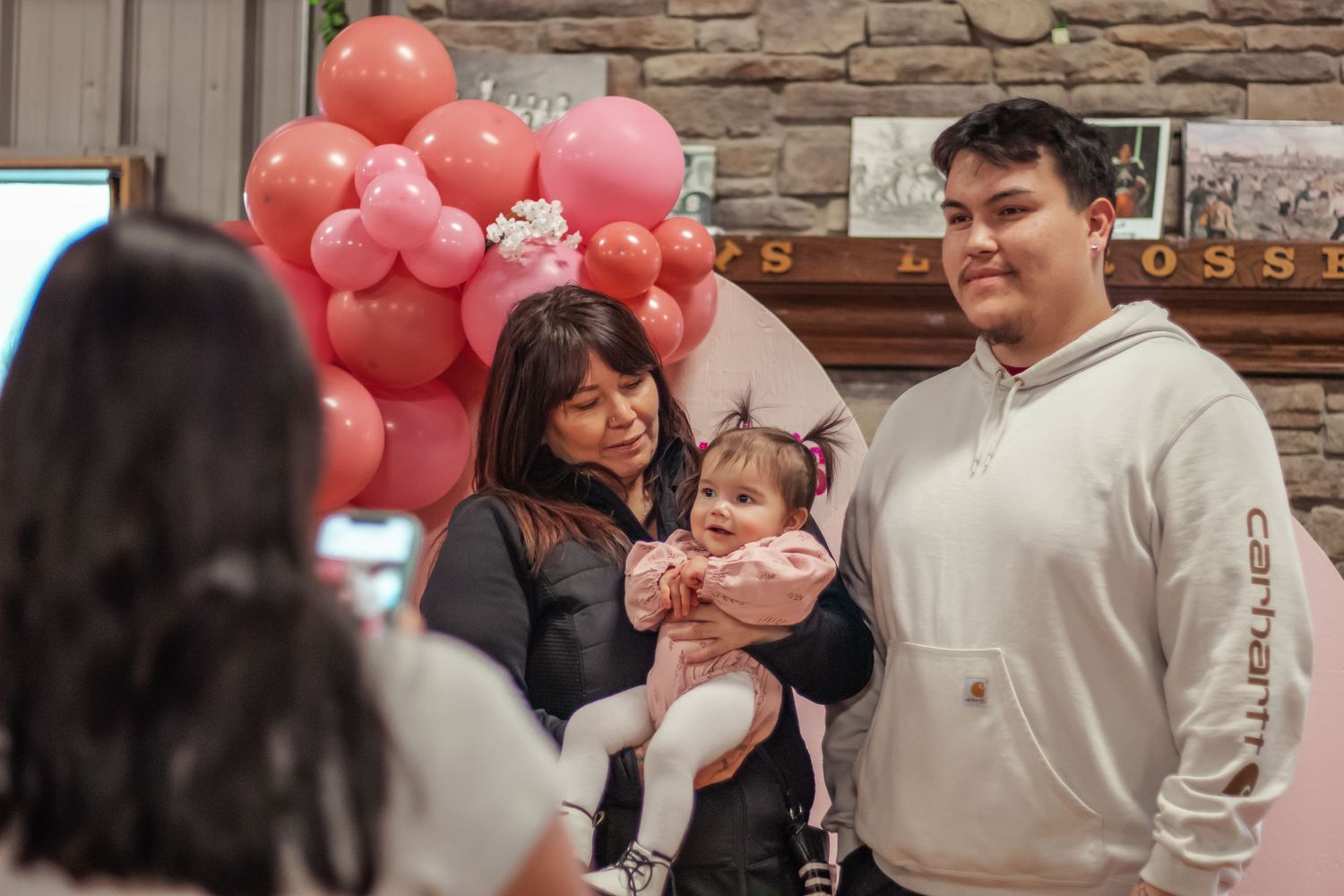 Family posing for photo at baby's first birthday party with pink balloon arrangement behind them.