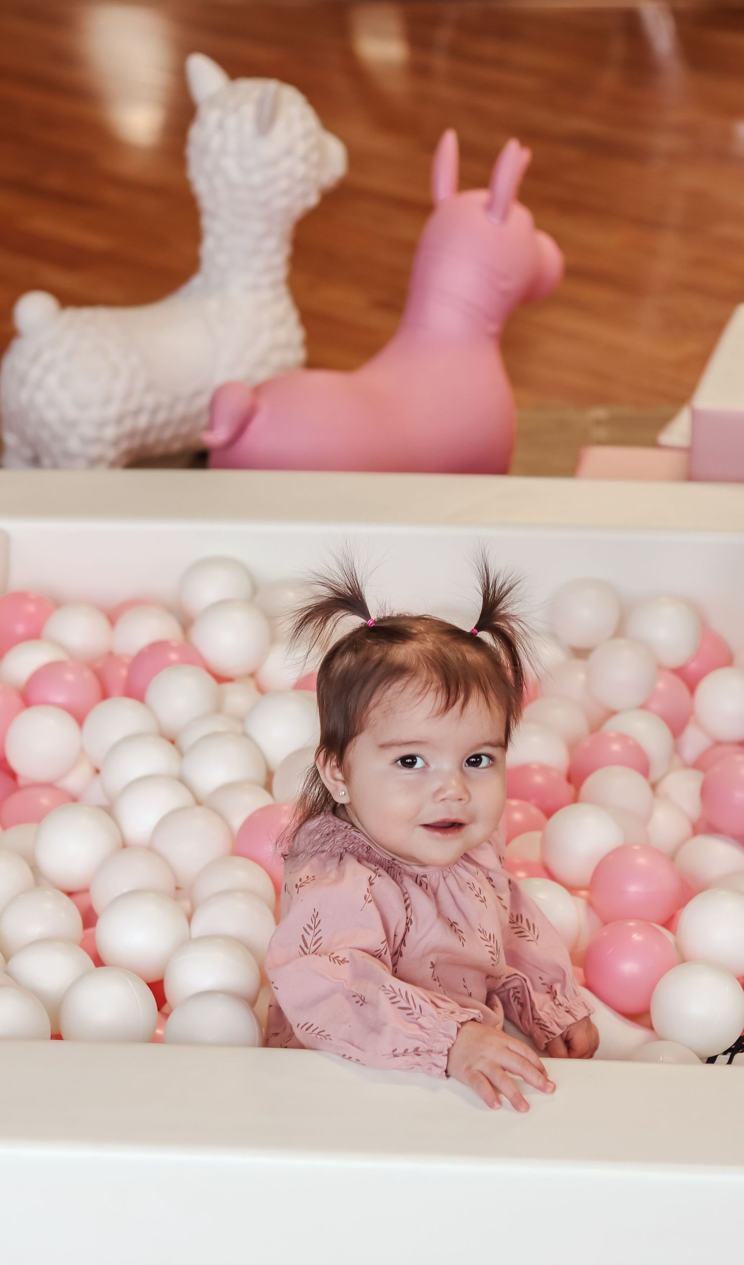 Baby girl in pink outfit sitting in a white and pink ball pit with toy llamas in the background.
